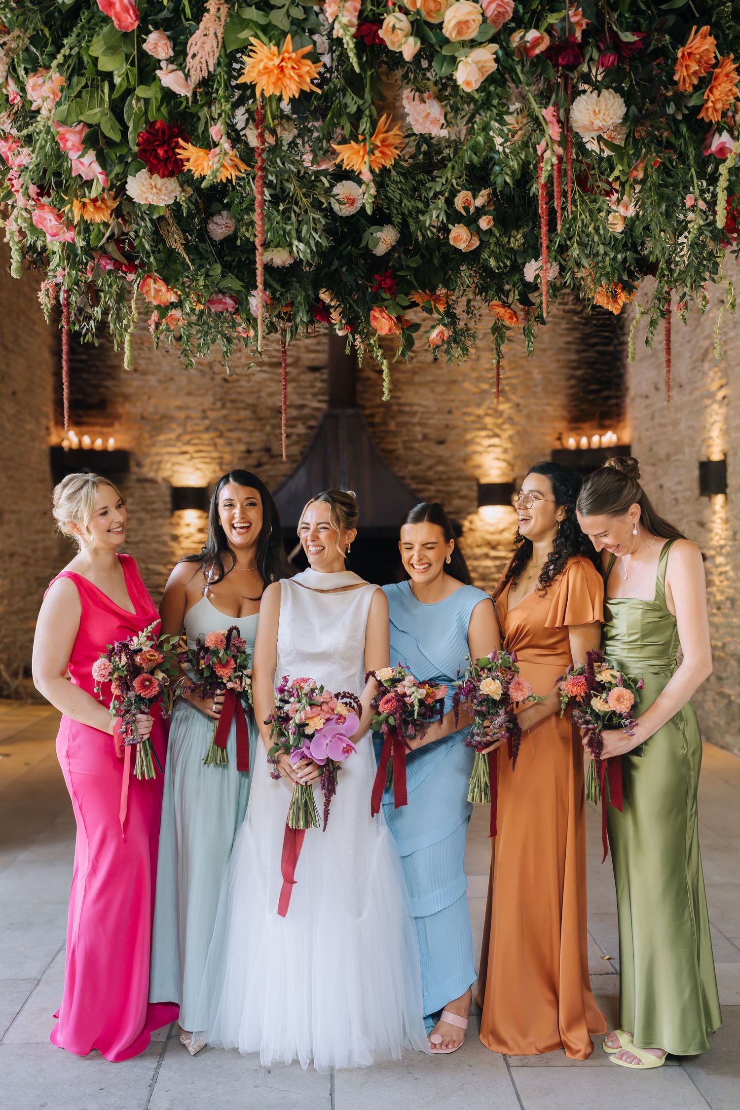Bride and five bridesmaids in colorful mismatched gowns pose under a lush floral installation at a wedding venue.