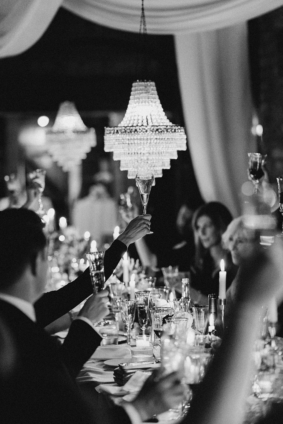 Guests raising champagne glasses in toast under crystal chandelier at elegant candlelit wedding reception.