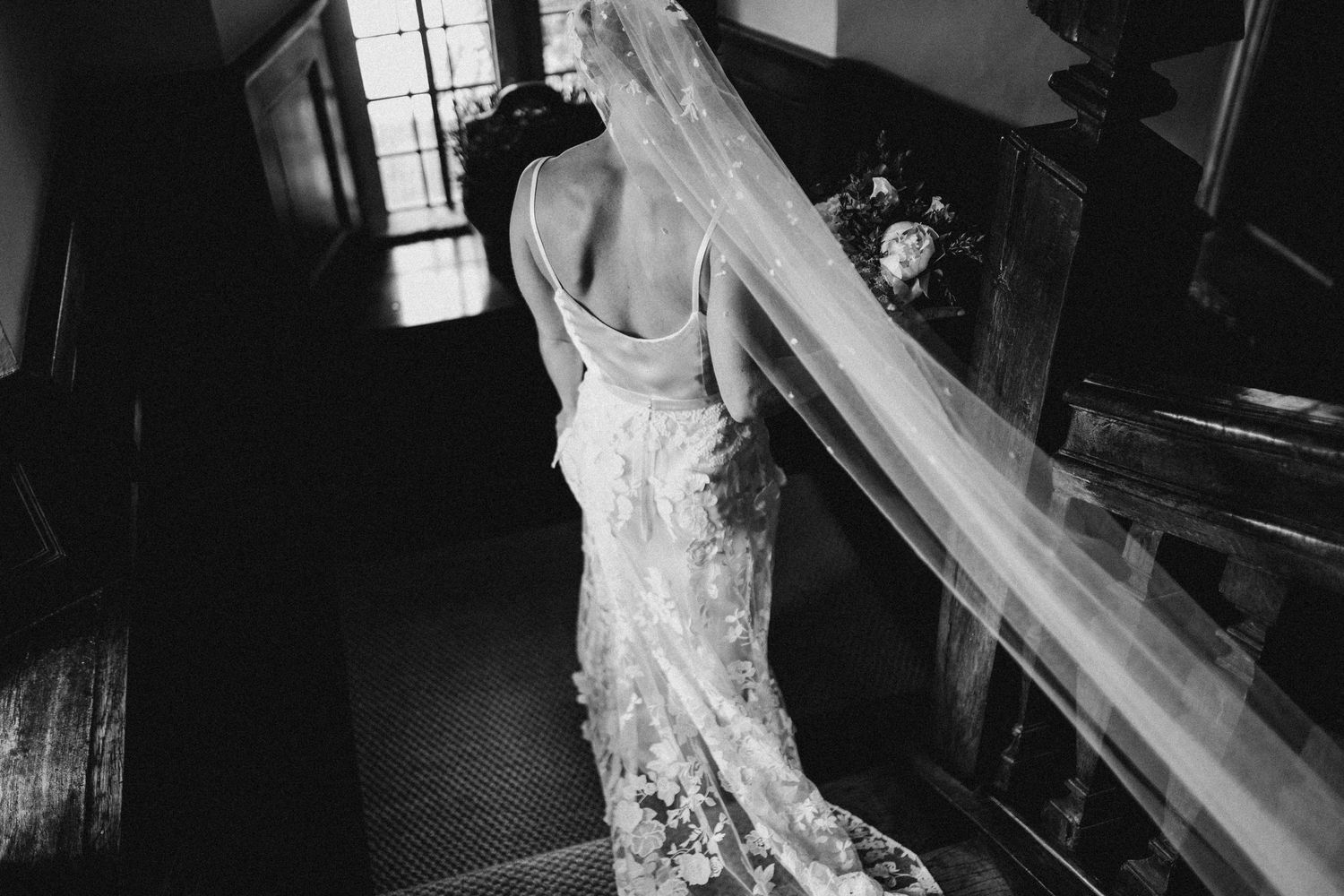 Black and white photo of bride in lace gown with veil descending elegant staircase with bouquet.