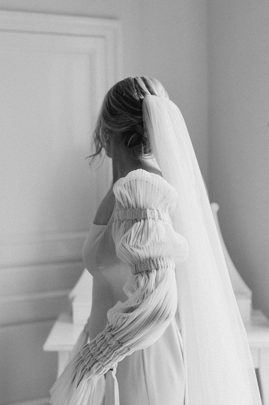 Elegant black and white rear view of bride in ruffled off-shoulder gown and long veil getting ready.