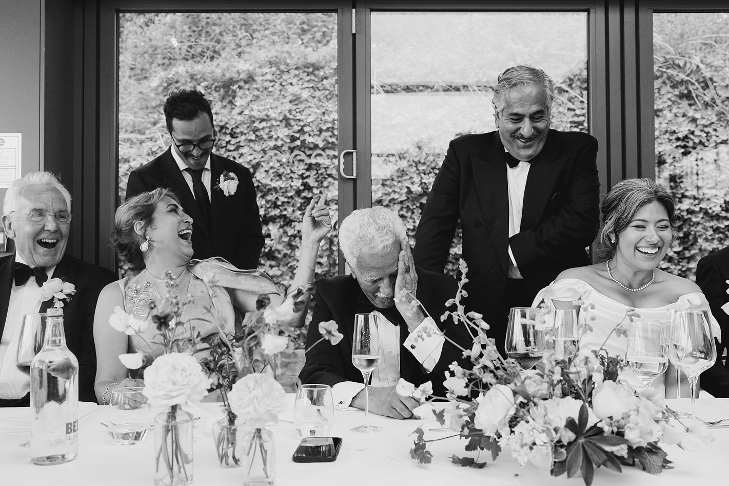 Wedding party laughing joyfully at the head table during reception speeches in a bright venue.