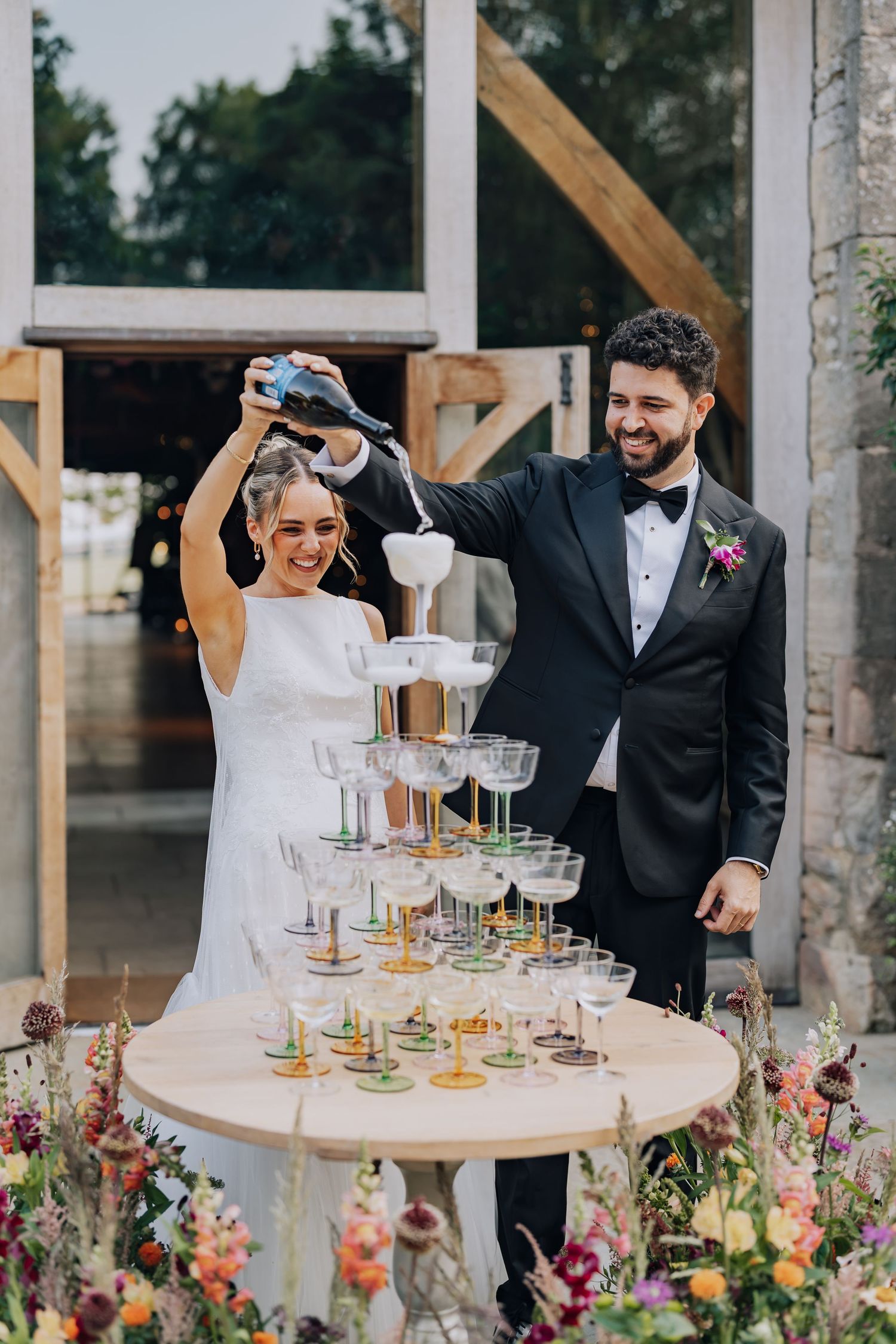 Bride and groom pouring champagne tower at wedding reception, surrounded by colorful floral arrangements.