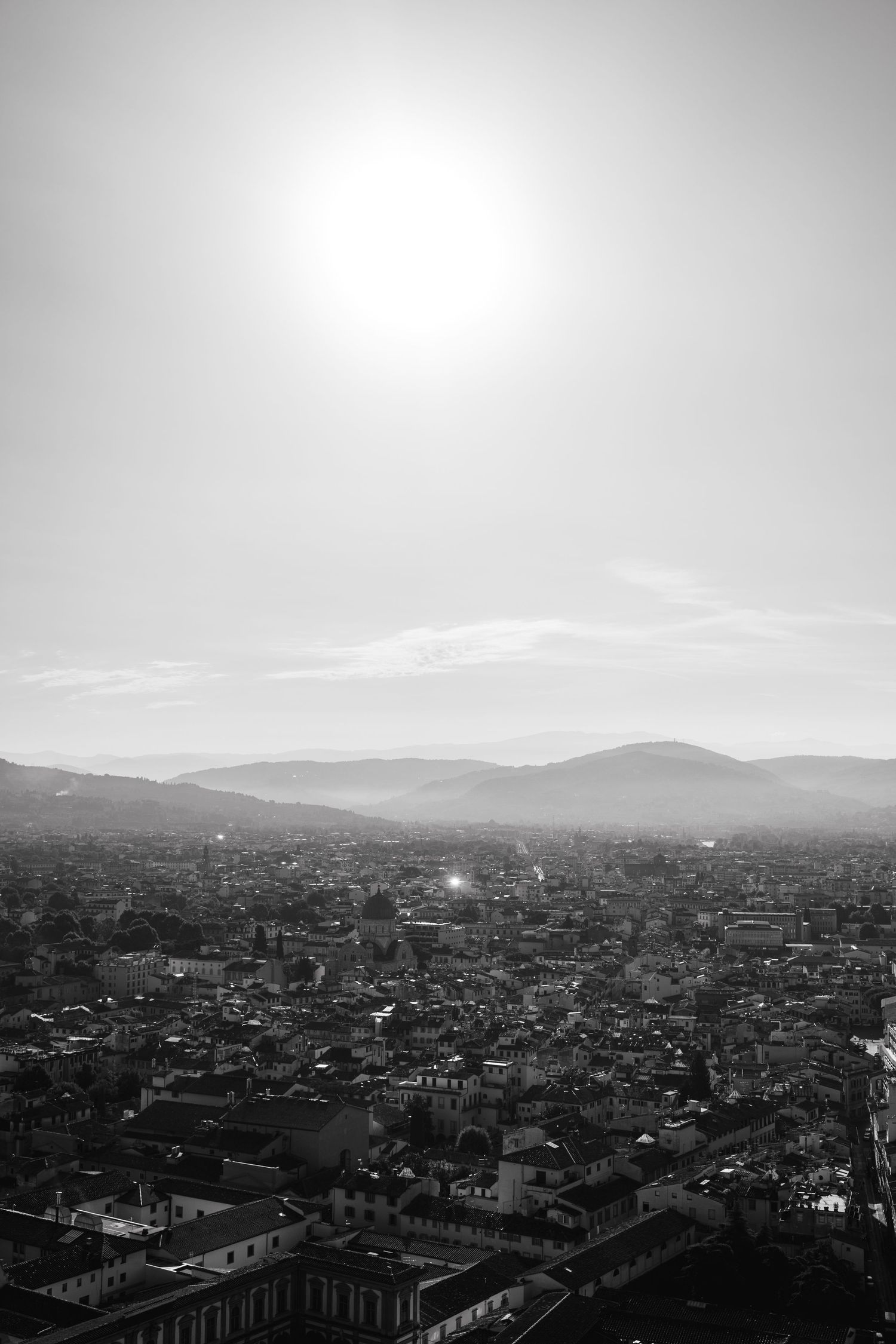 Black and white aerial cityscape with sun glare over rooftops and distant mountains.