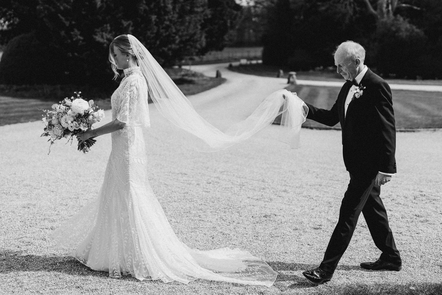 Black and white photo of a bride in a lace gown and veil walking with an older gentleman in a tuxedo.