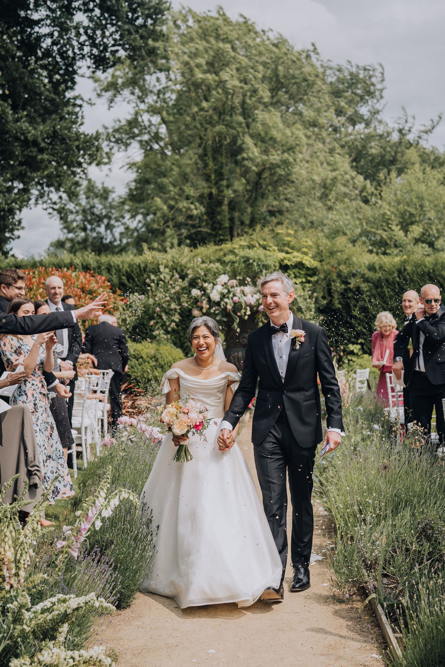 Smiling bride and groom walk hand-in-hand down lavender-lined aisle at outdoor garden wedding.