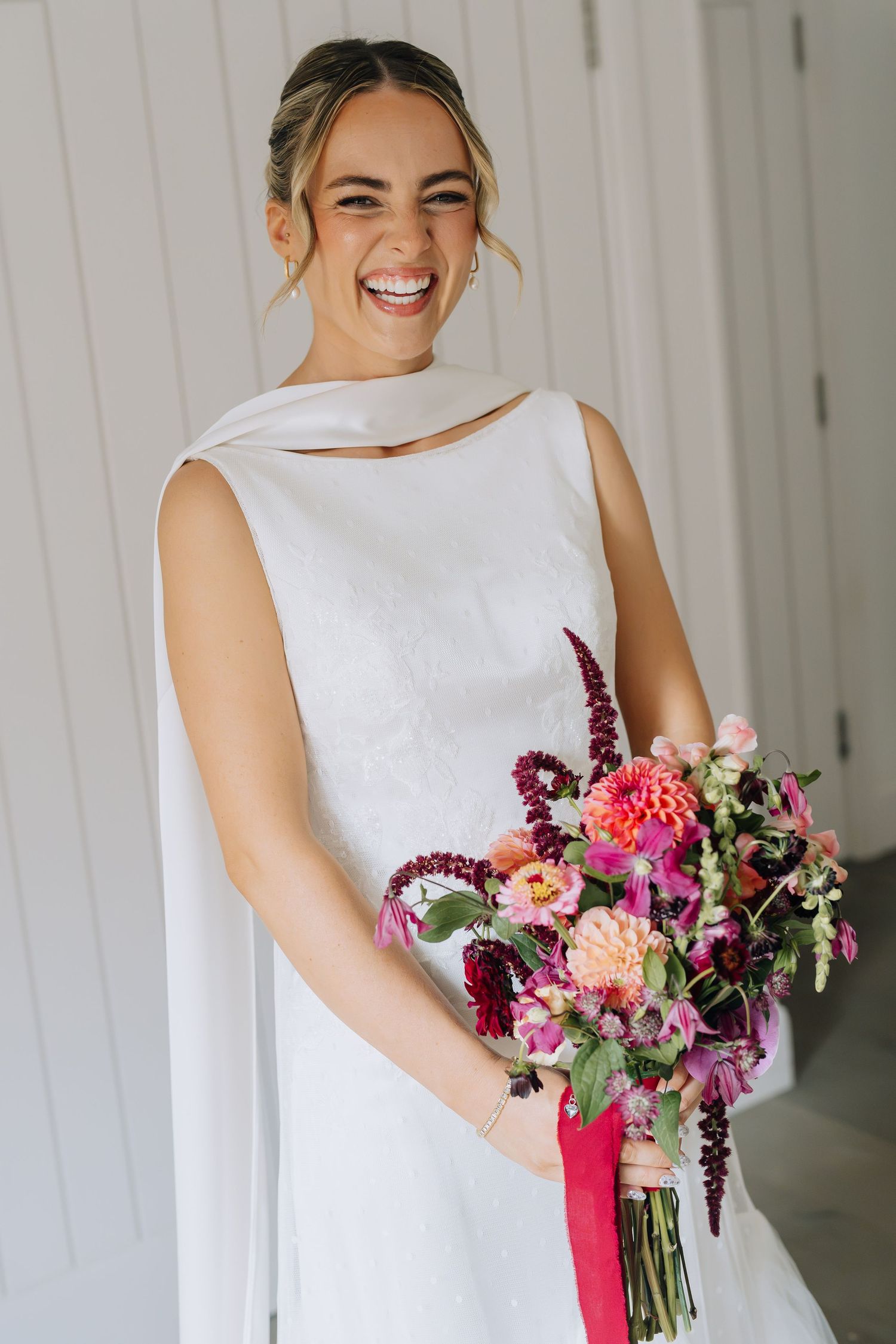 Smiling bride in white sleeveless gown holding vibrant pink and burgundy wedding bouquet with ribbon.