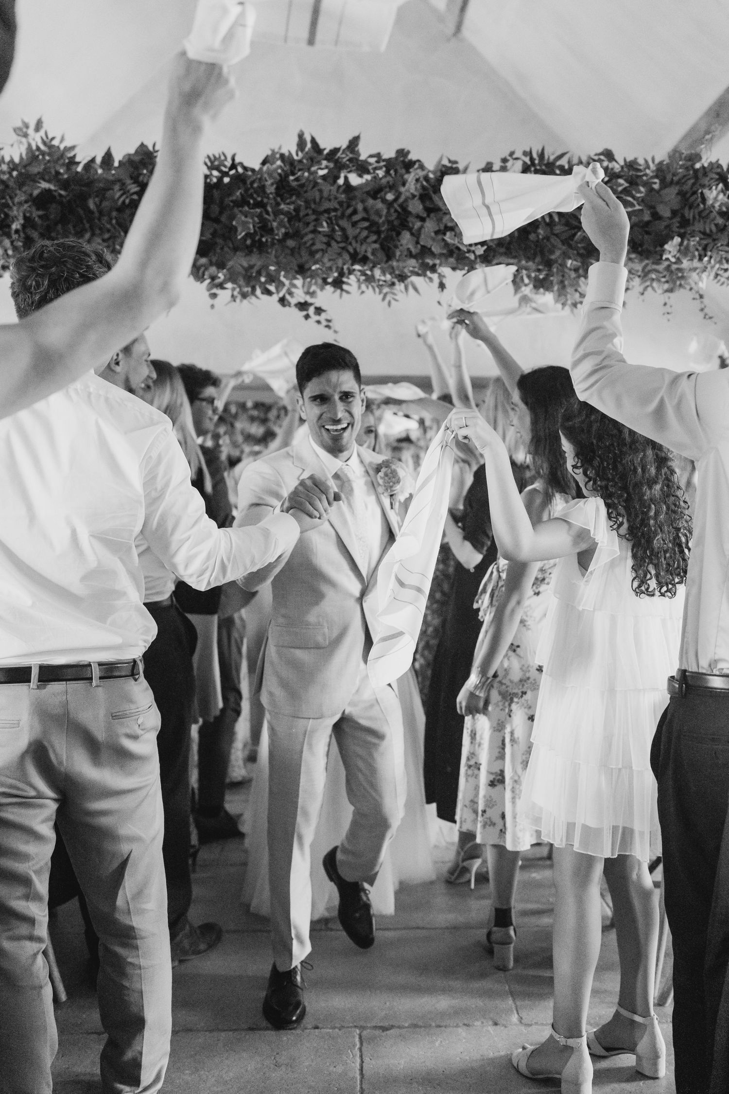 Groom in white suit dancing joyfully with wedding guests waving napkins during celebratory reception exit.