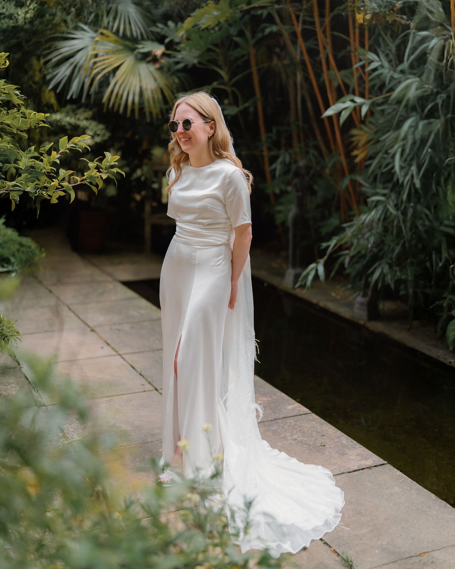 Bride in white satin two-piece wedding dress with train, wearing sunglasses, posing in lush garden setting.