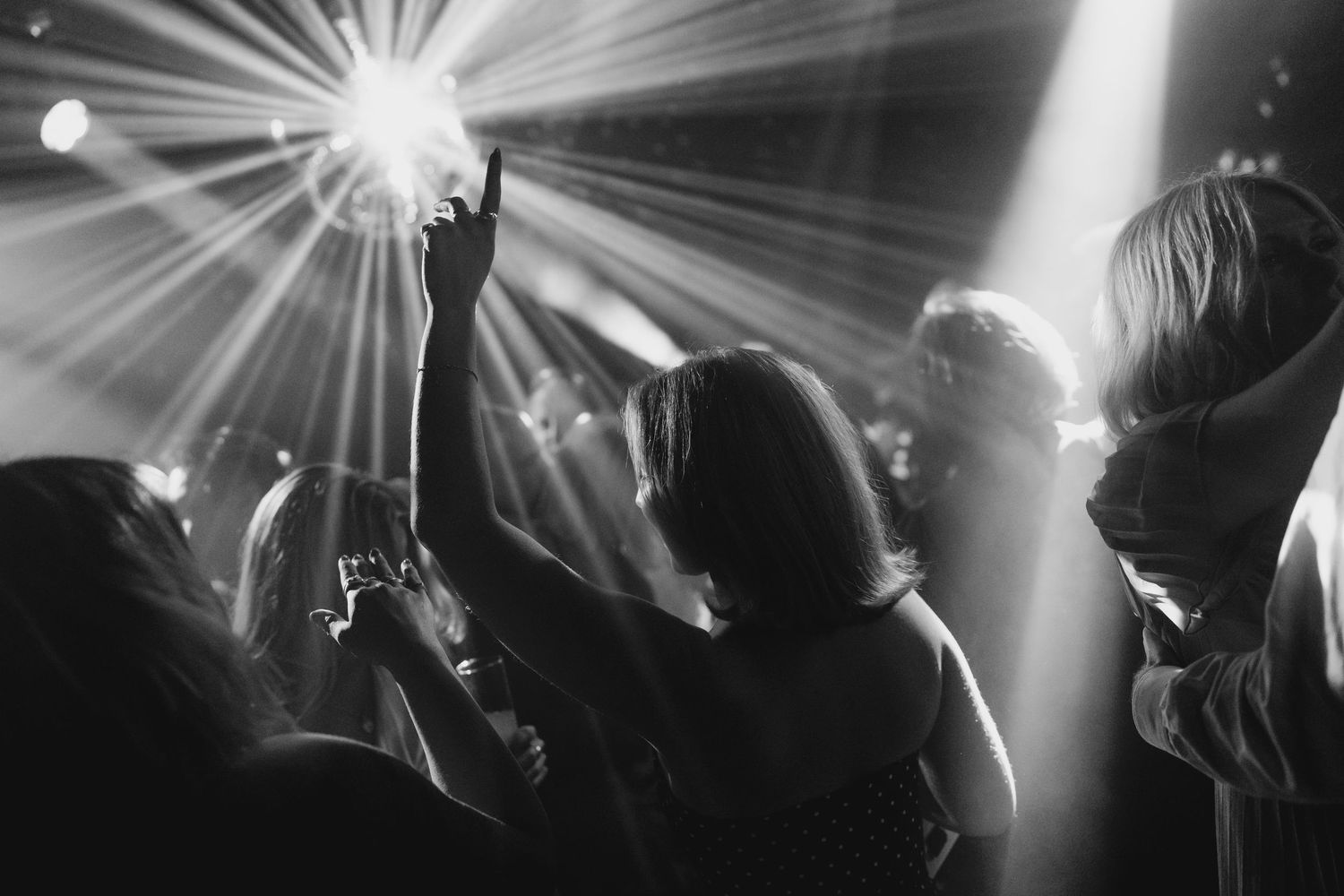 Black and white photo of people dancing at a nightclub with dramatic stage lighting and raised hands.
