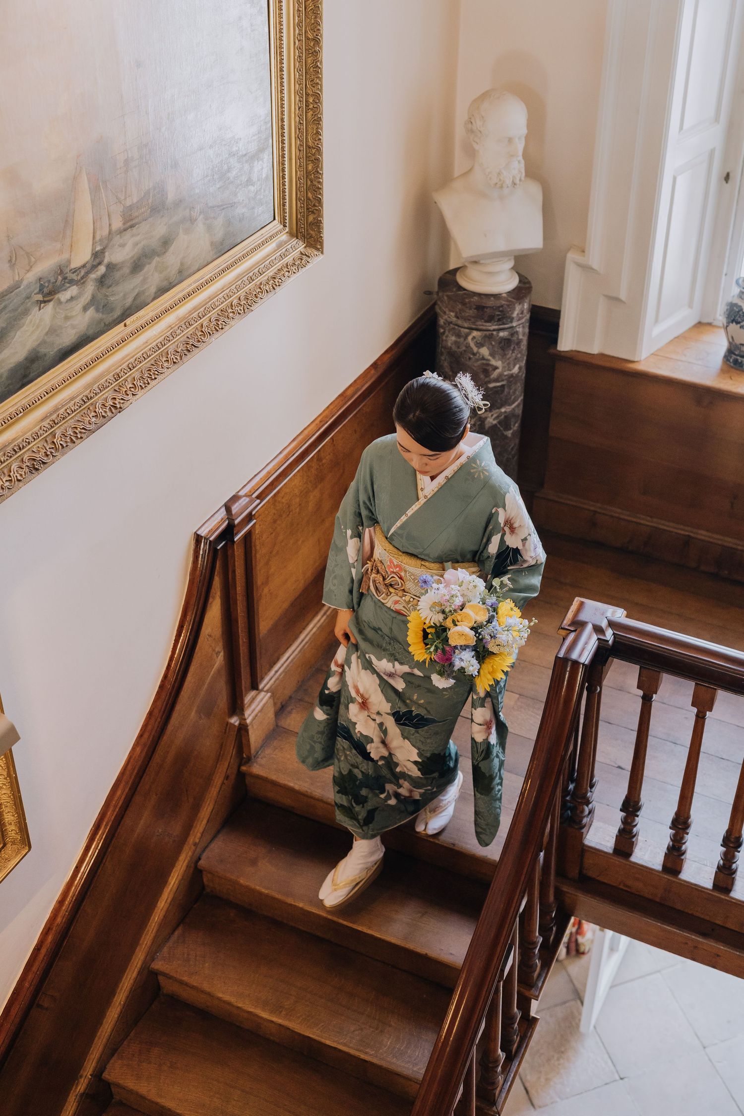 Woman in green kimono holding bouquet descends wooden staircase in elegant interior with painting and marble bust.