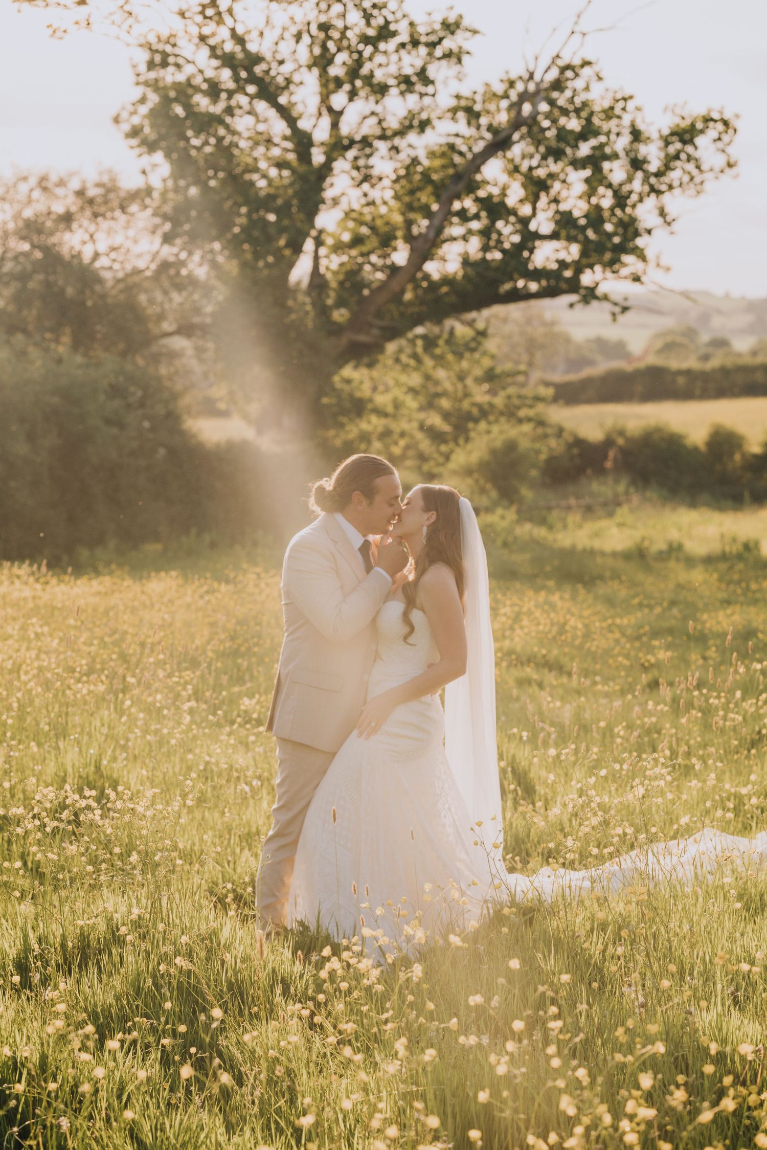 Bride and groom sharing golden hour kiss in sun-drenched wildflower meadow with veil flowing.