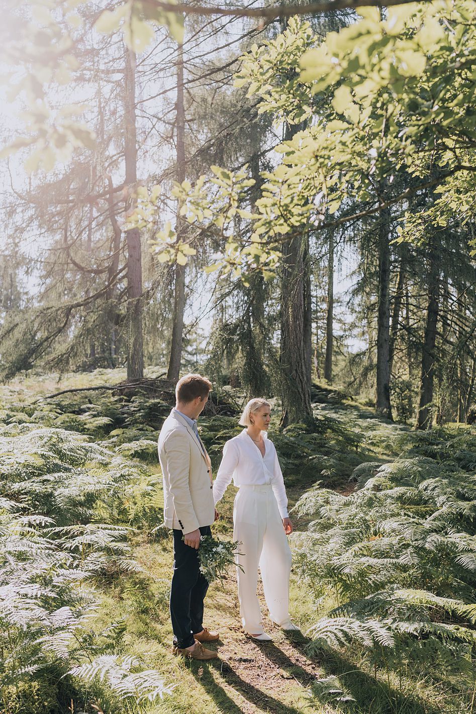 Bride and groom holding hands walking through a sunlit forest with lush green ferns.