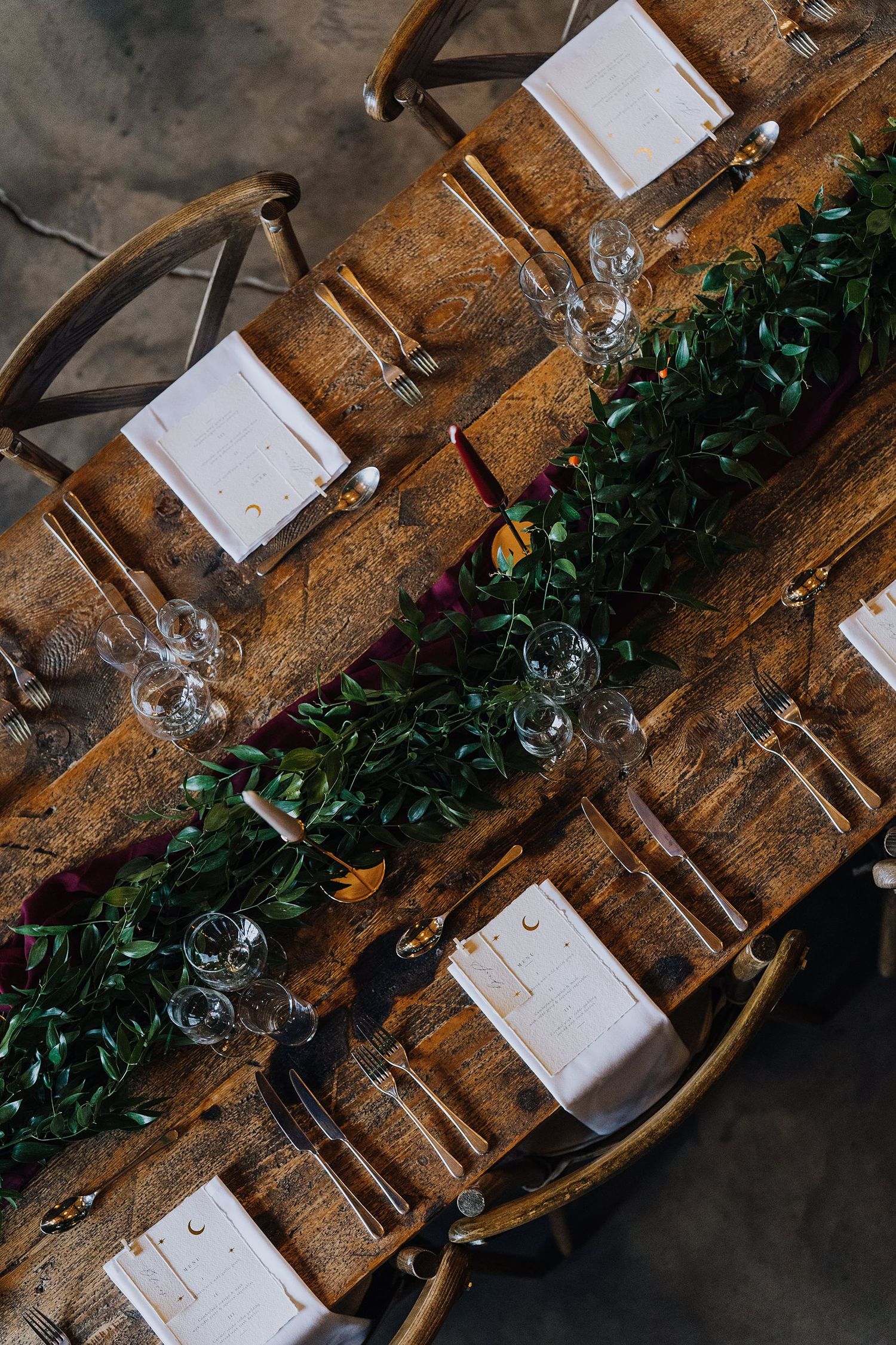 Overhead view of a rustic wooden reception table set with greenery runner and white menu cards.
