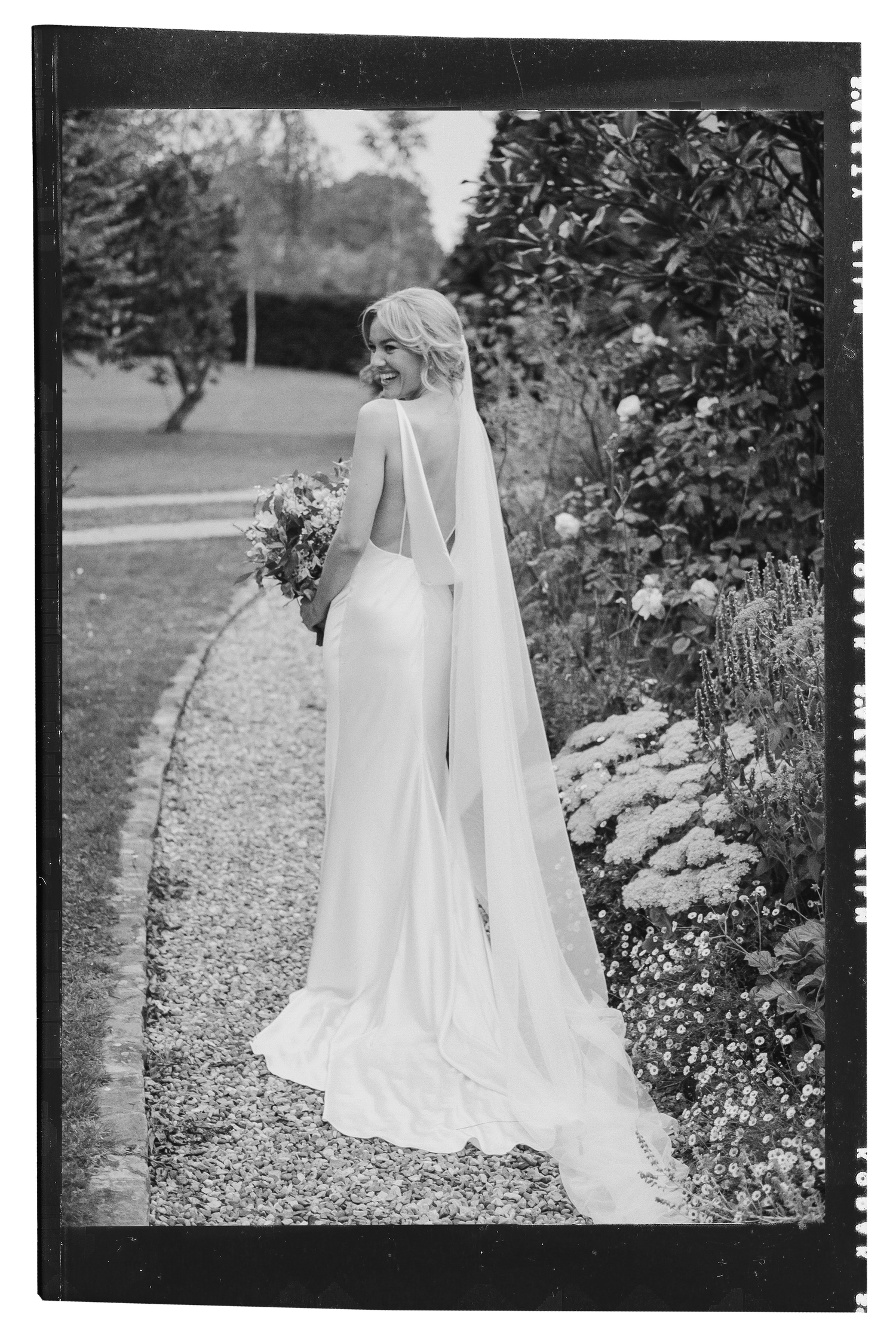 Bride in backless white wedding gown with veil walking along garden path, black and white photo.