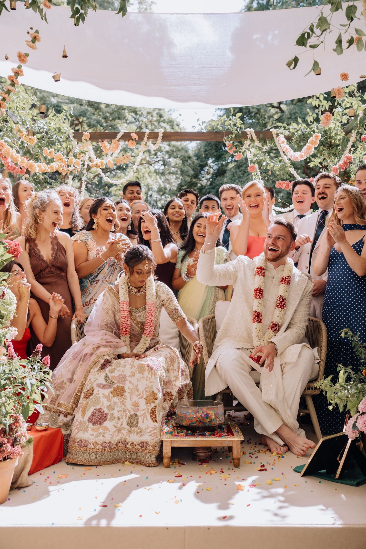 Joyful Indian wedding ceremony with bride in embroidered lehenga and groom in white sherwani surrounded by cheering guests outdoors.