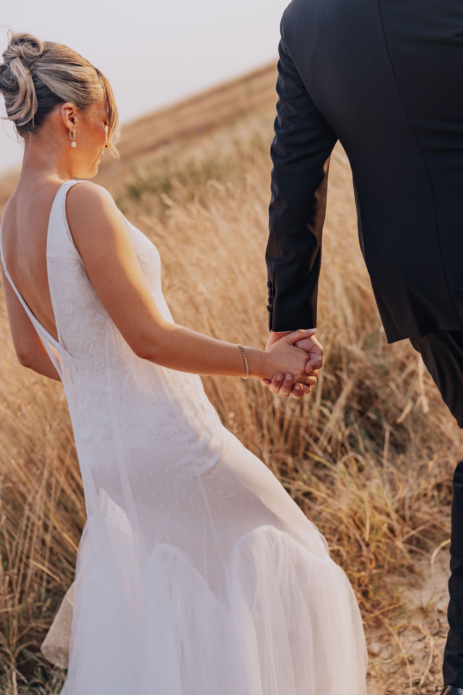 Bride in white backless gown holds groom's hand in golden field at sunset during outdoor wedding.