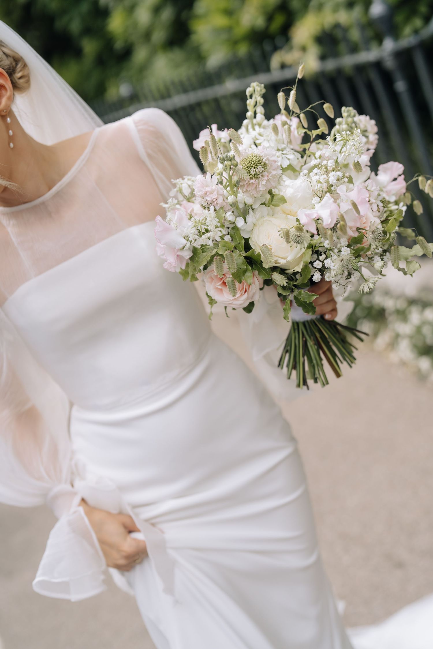 Bride in white satin gown and veil holding elegant blush and white floral bouquet with greenery outdoors.
