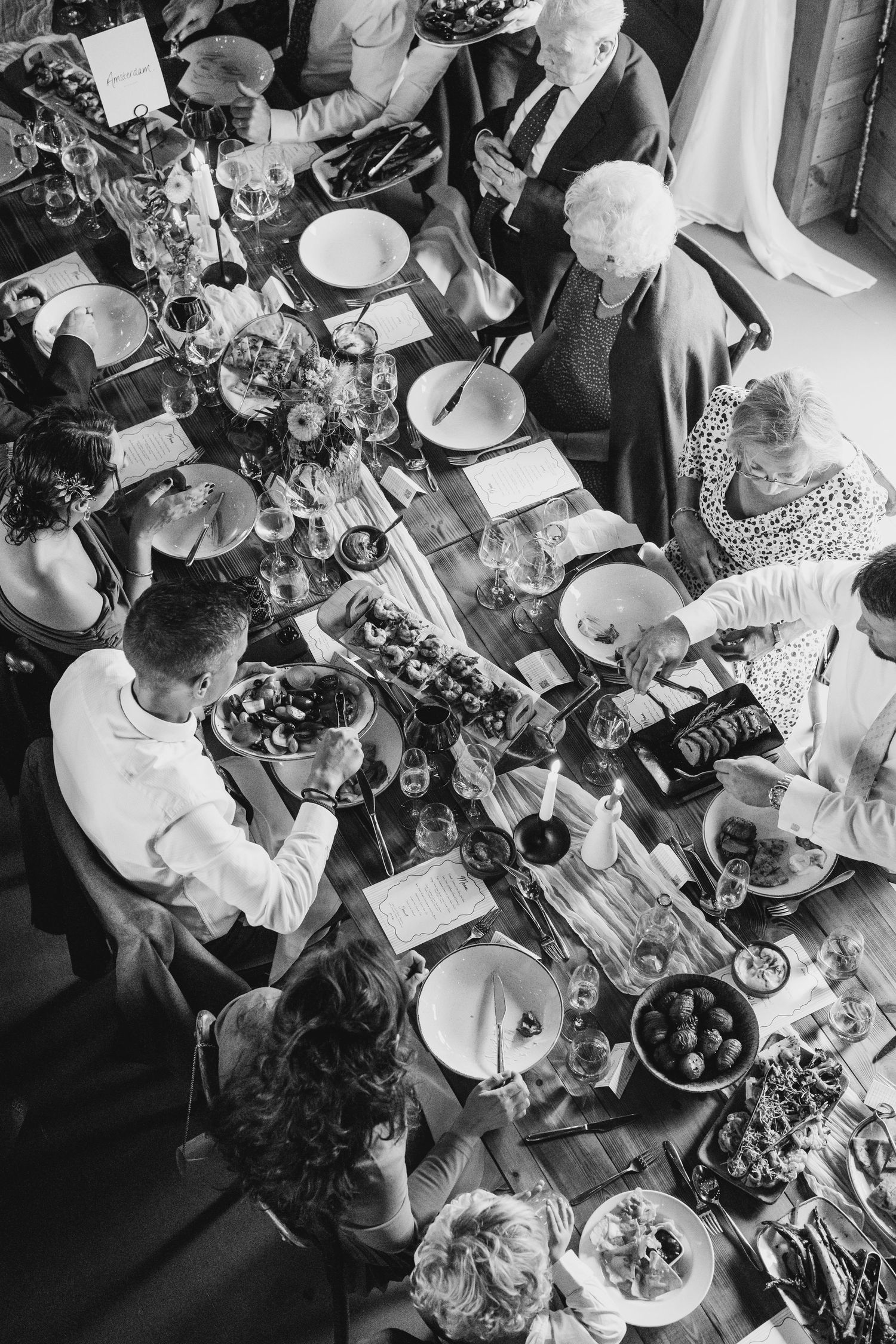Aerial black and white view of wedding guests dining at a beautifully decorated long reception table.