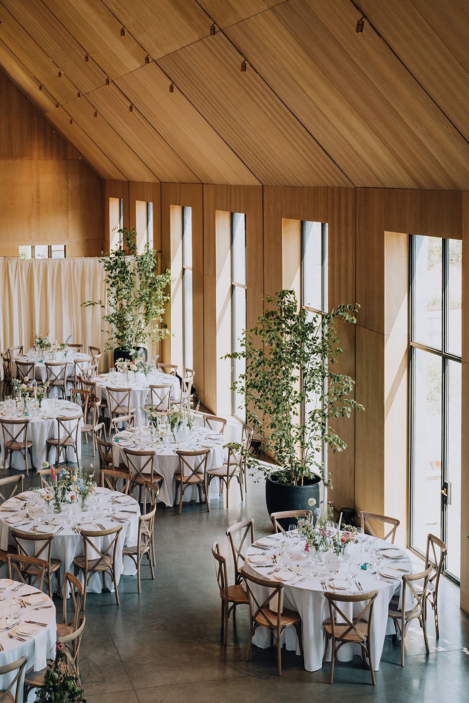 Bright modern wedding reception venue with wooden ceiling, tall greenery centerpieces and round tables with white linens.