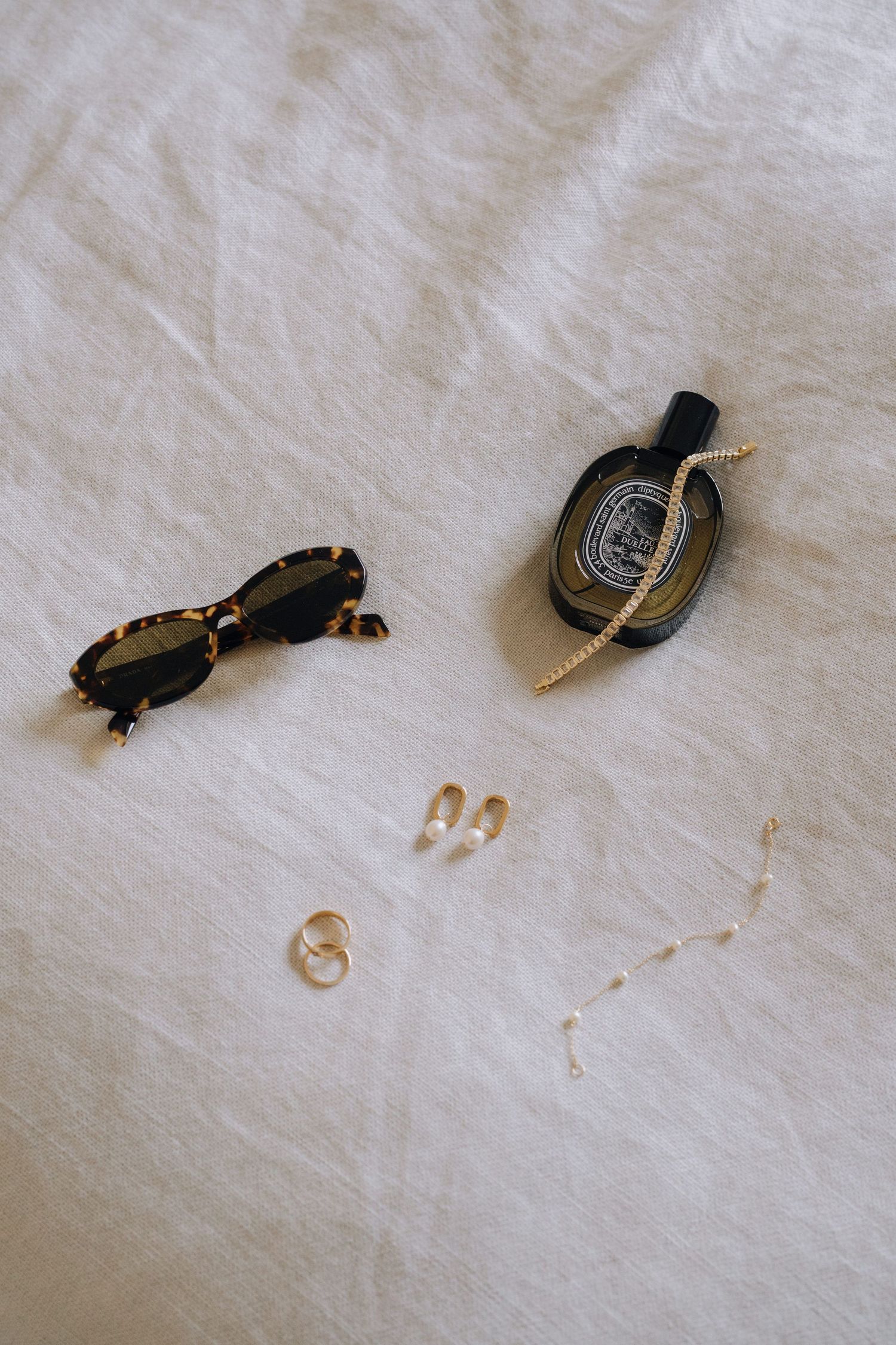 Flat lay of tortoiseshell sunglasses, Diptyque perfume, gold rings, pearl earrings, and delicate chain on white linen.