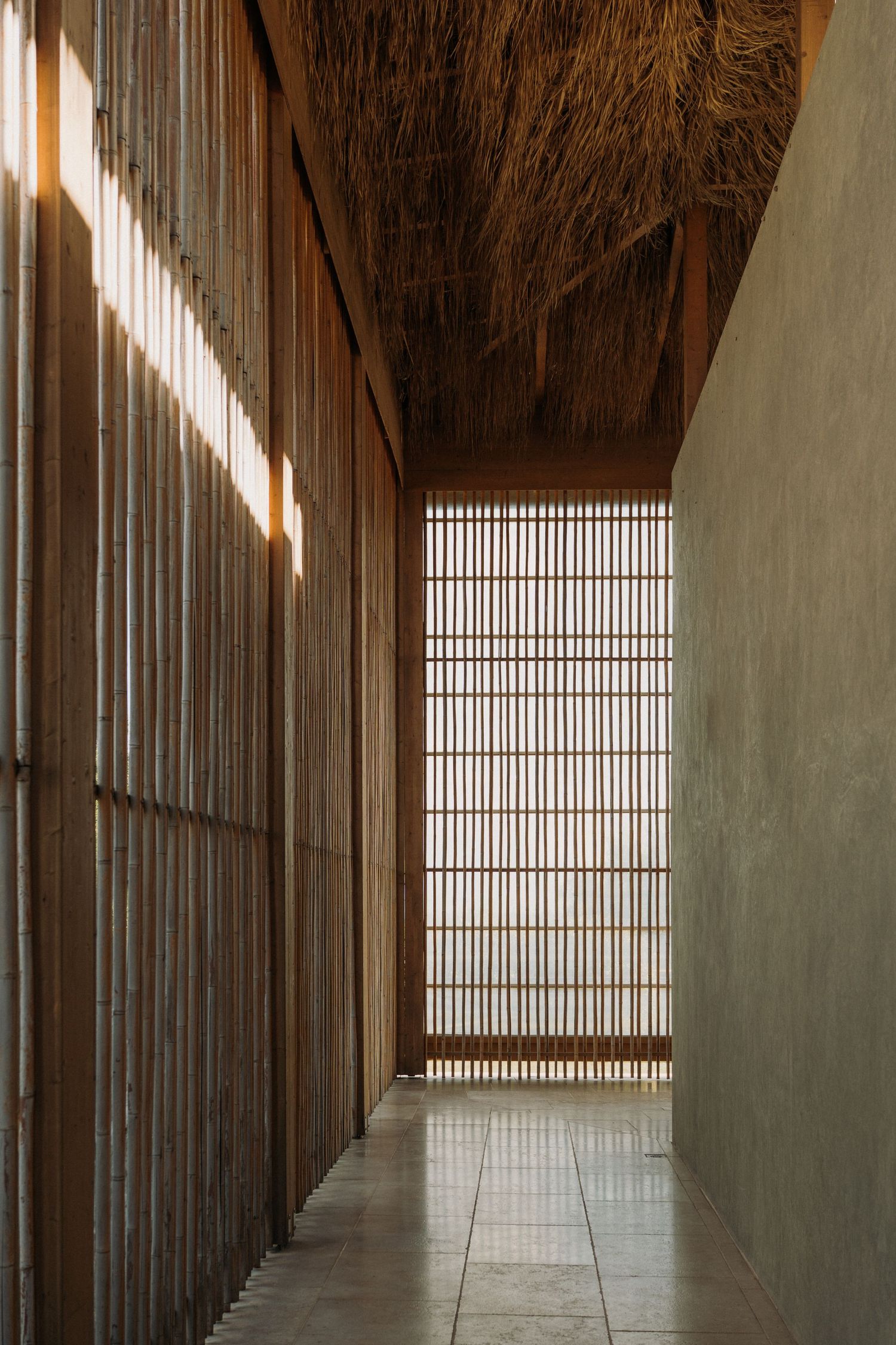 Narrow corridor with bamboo slat walls, thatched ceiling, and natural light in tropical architecture.