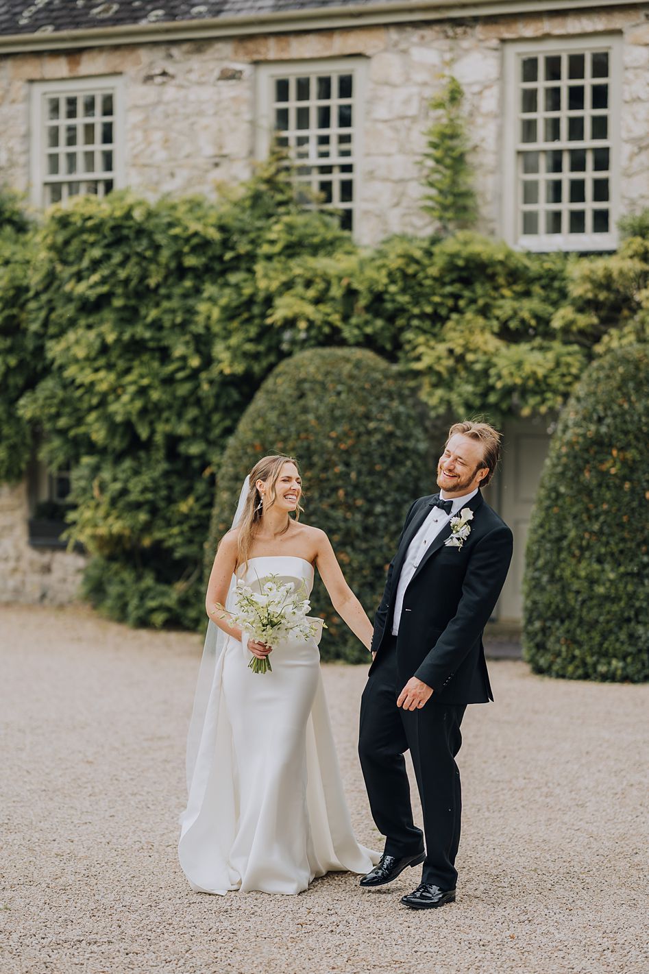 Bride in strapless white gown and groom in black tuxedo standing before an ivy-covered stone estate.
