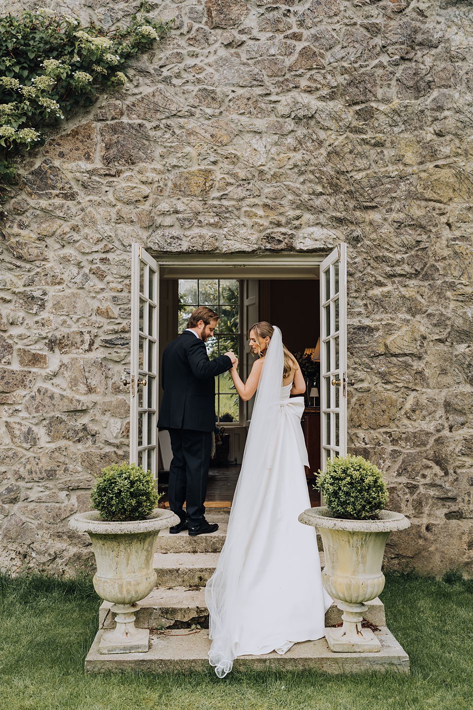 Bride and groom share an intimate moment in doorway of rustic stone building flanked by topiary urns.