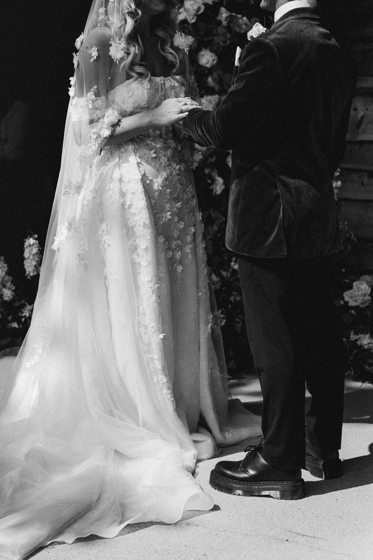 Black and white photo of bride in floral lace gown and groom in velvet suit standing at wedding altar.