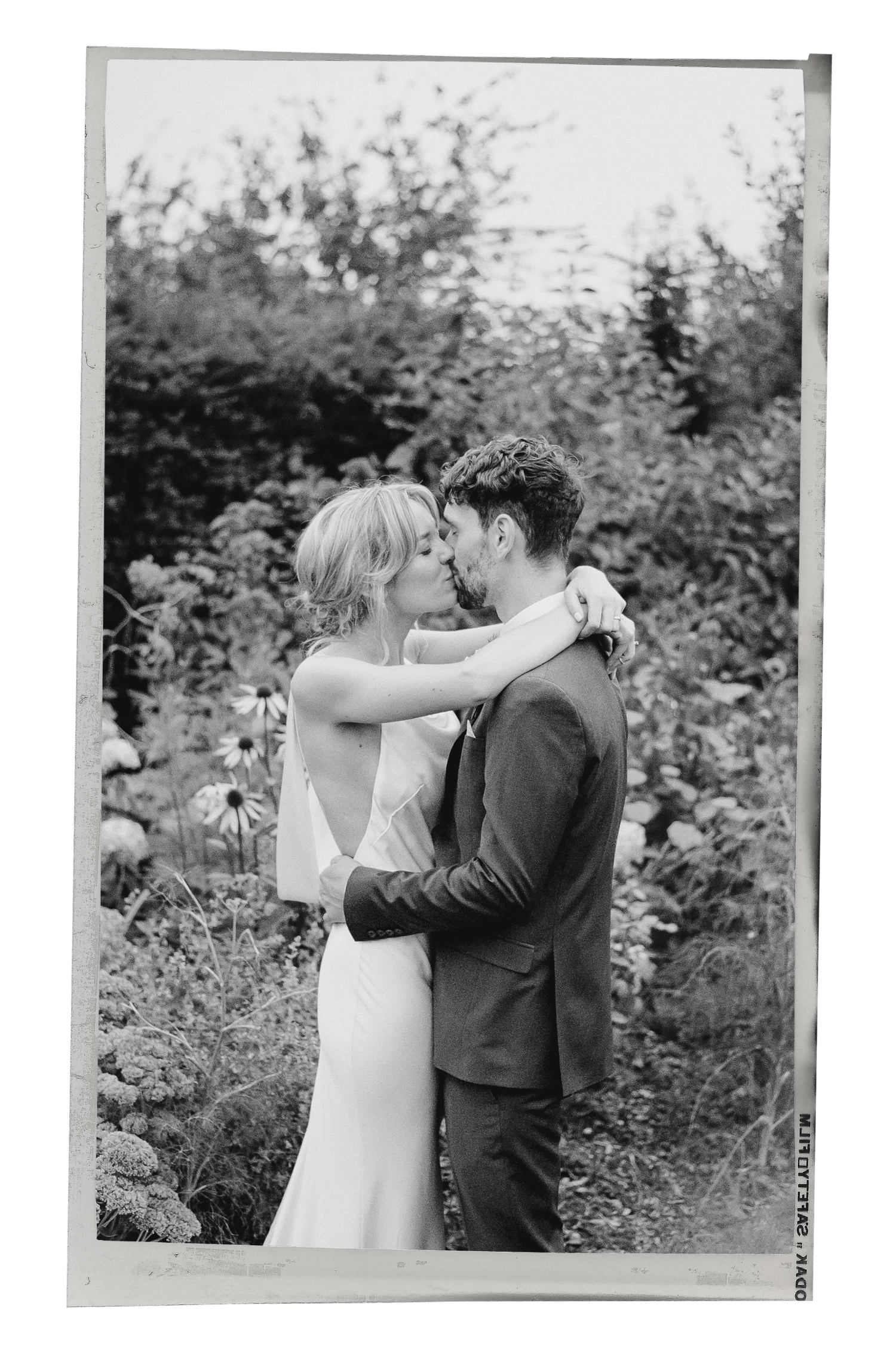 Black and white film photo of bride and groom kissing in a lush garden setting.
