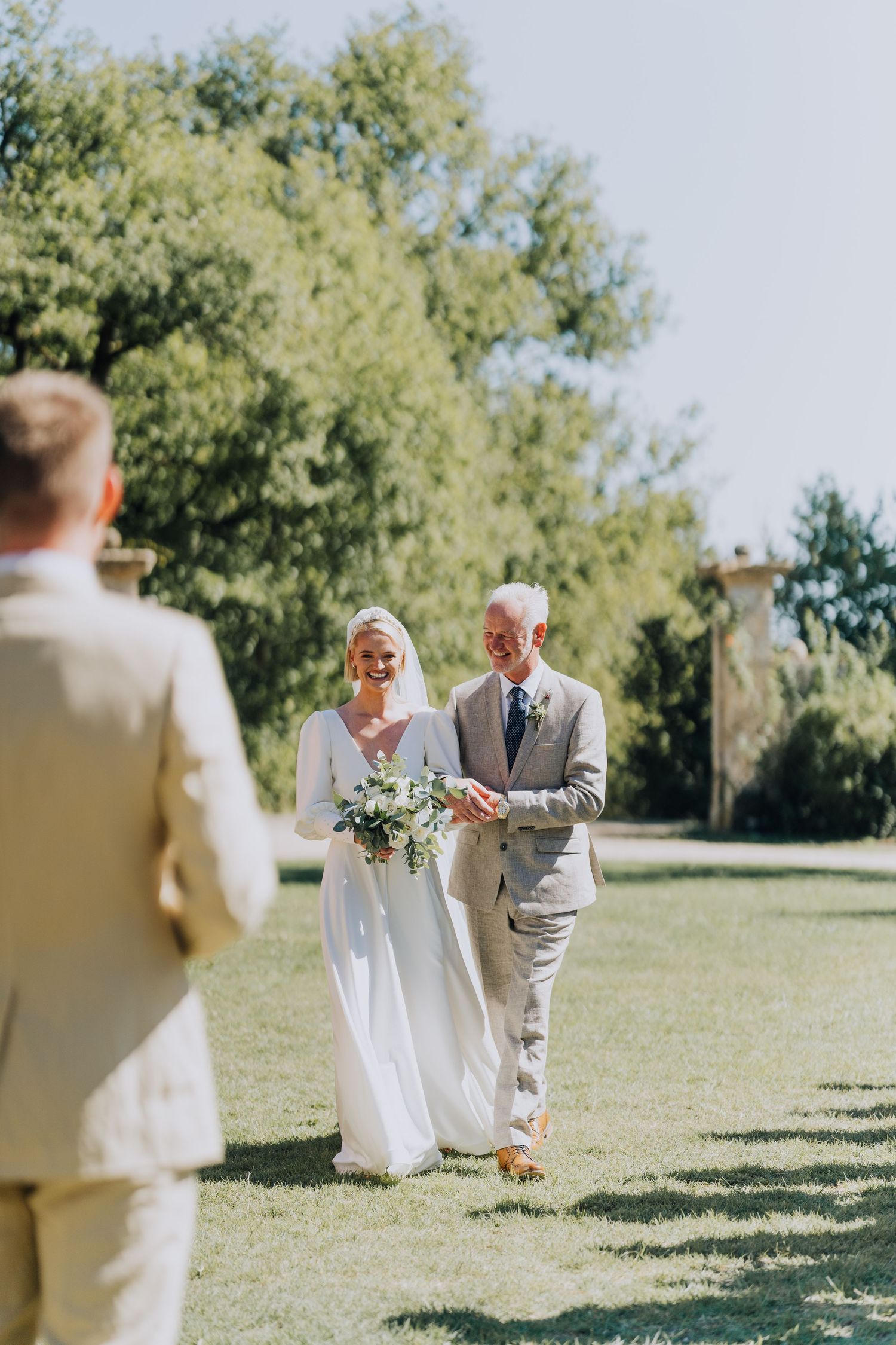 Bride walking down outdoor aisle with father toward groom at sunny garden wedding.