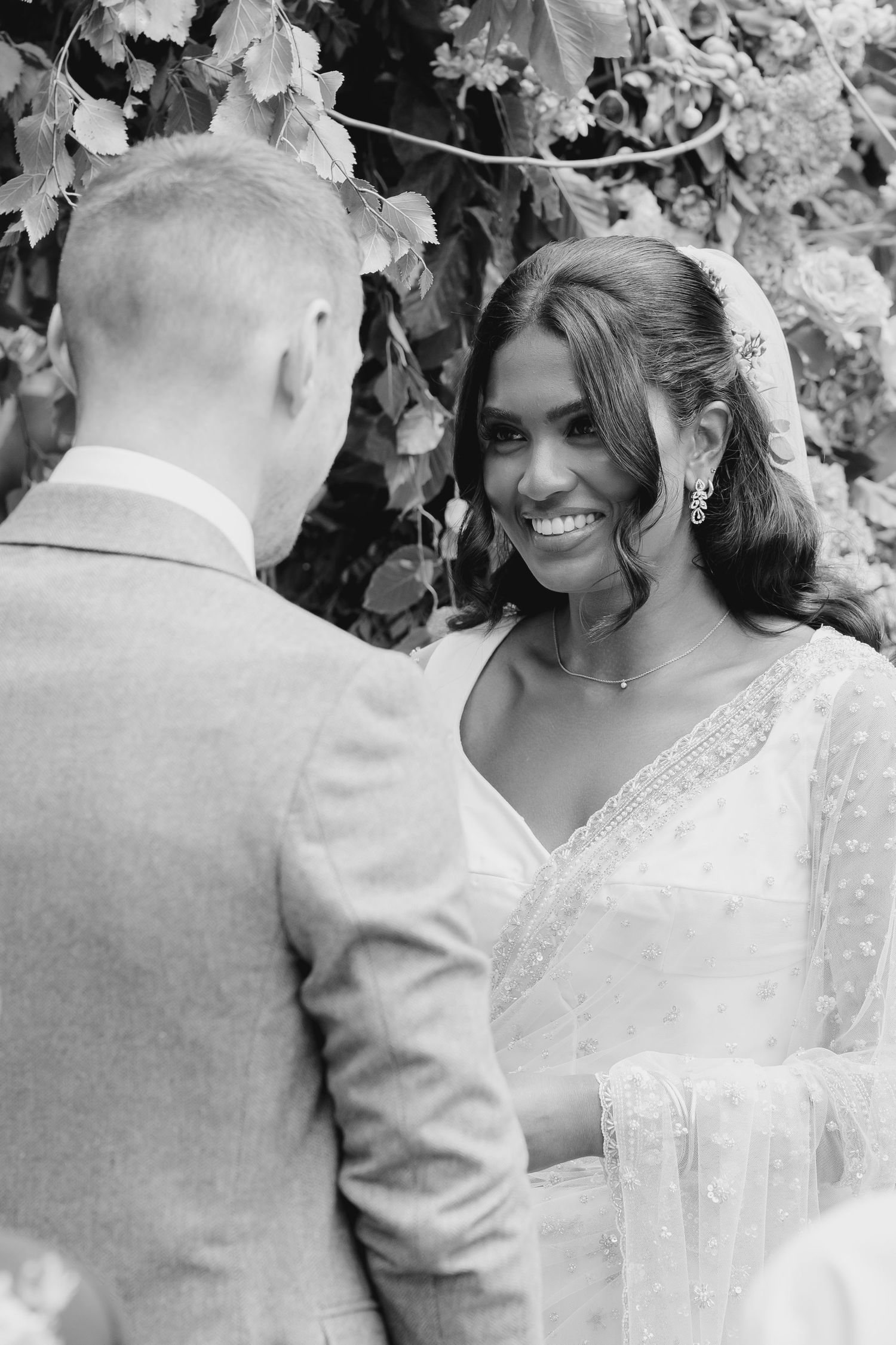 Black and white photo of radiant bride smiling at groom during outdoor wedding ceremony beneath floral arch.