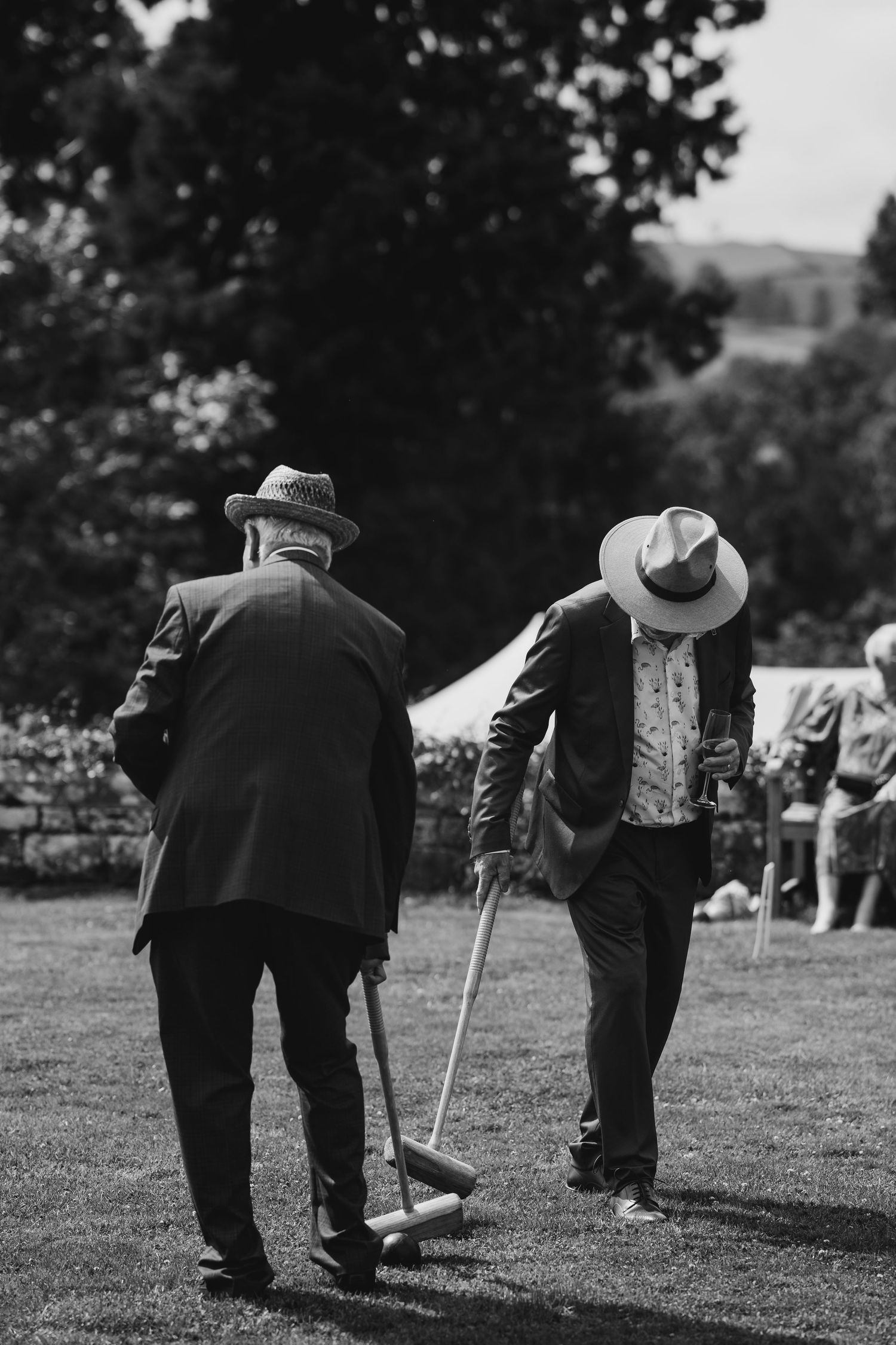 Two elderly men wearing straw hats playing croquet on lawn at outdoor wedding celebration.