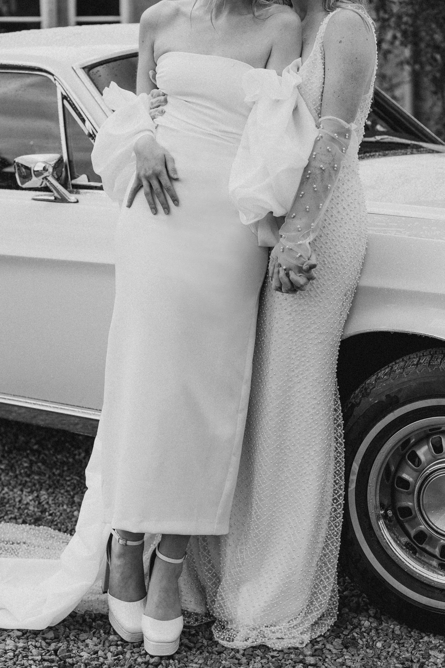 Two brides in elegant white gowns pose together beside a classic white car in a black and white wedding photo.