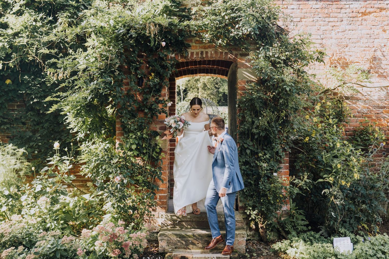 Bride in white gown and child in blue suit standing beneath a flower-covered brick archway.