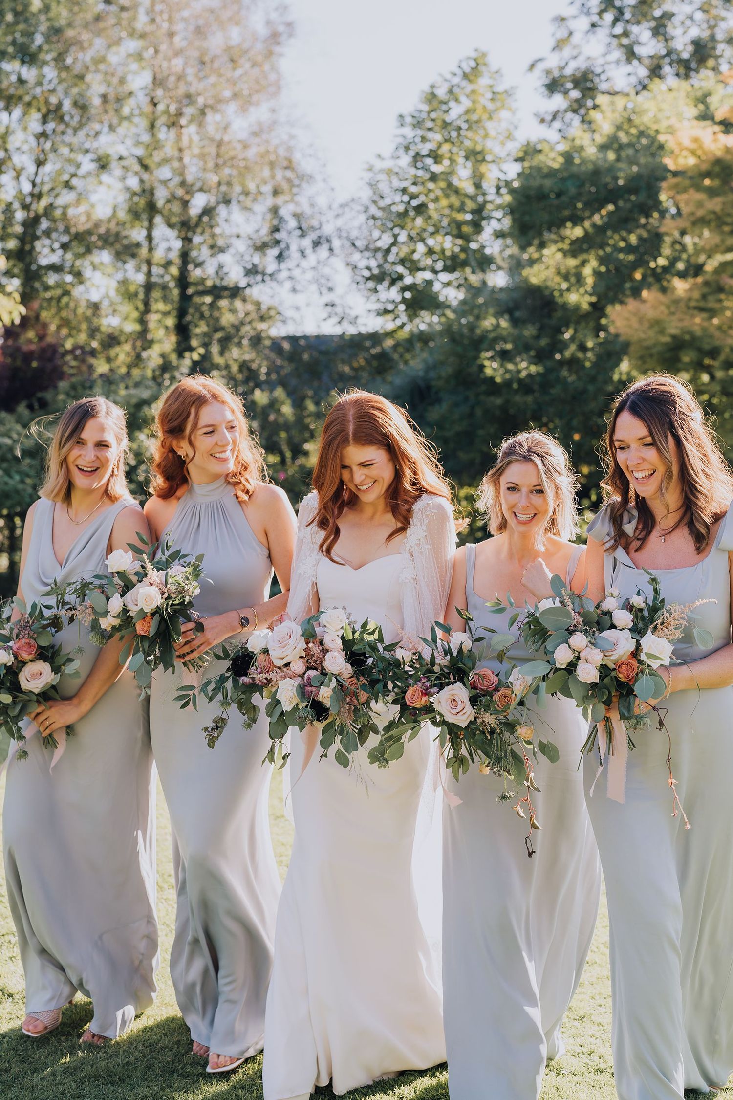 Bride and four bridesmaids in sage green dresses walk outdoors laughing with floral bouquets.