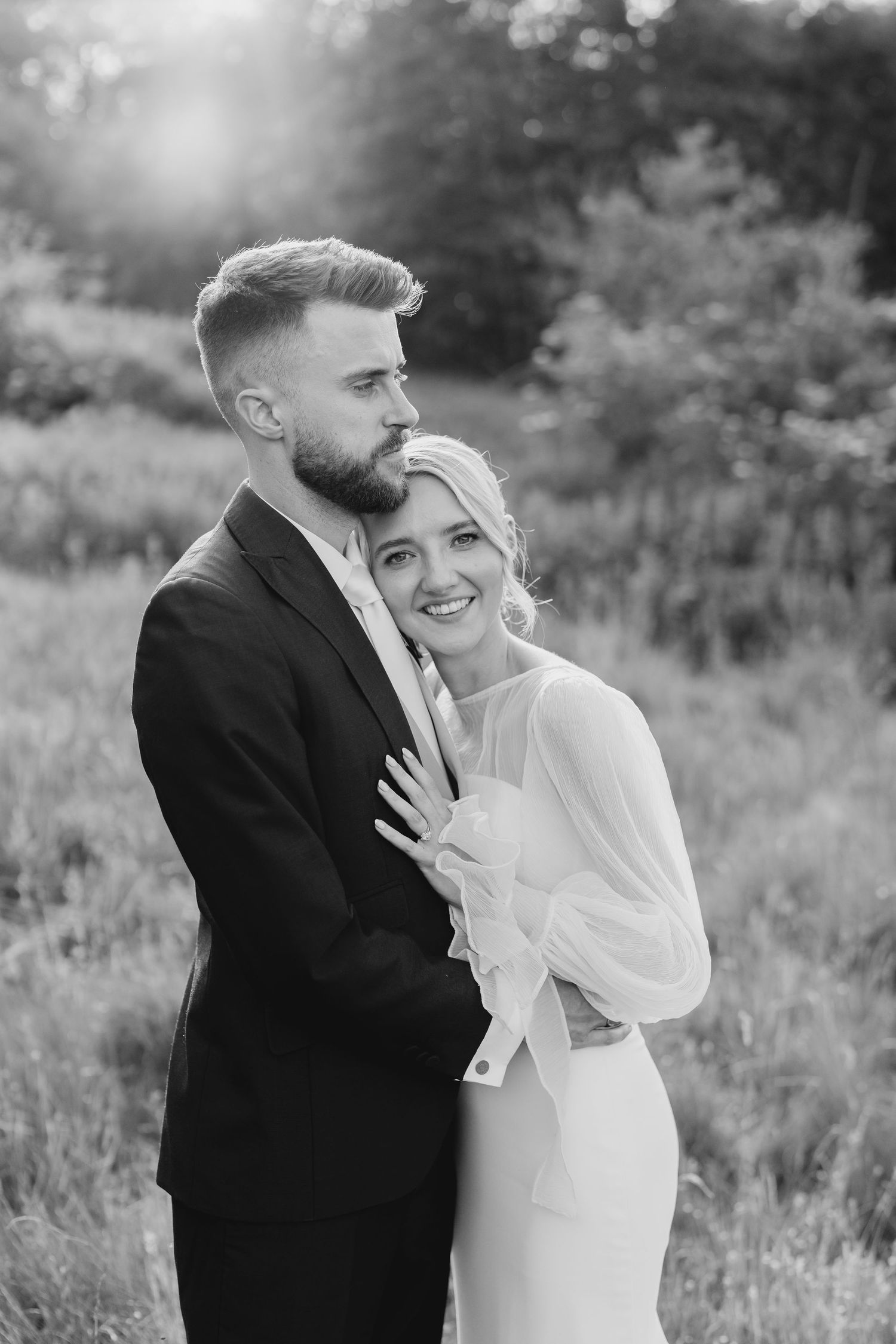 Black and white photo of a smiling bride in a white gown embracing a bearded groom in a suit outdoors.