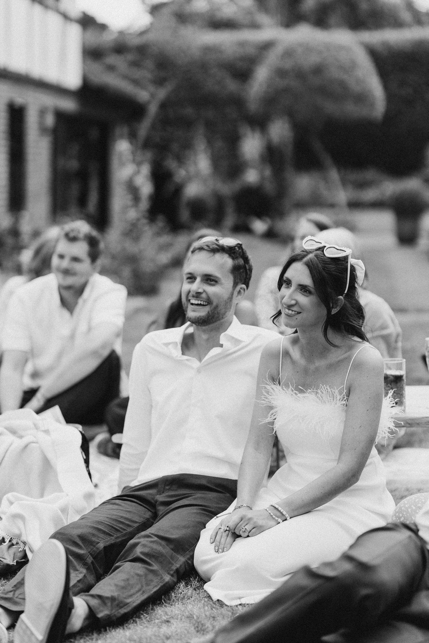 Black and white photo of smiling wedding guests relaxing on grass during an outdoor summer reception.