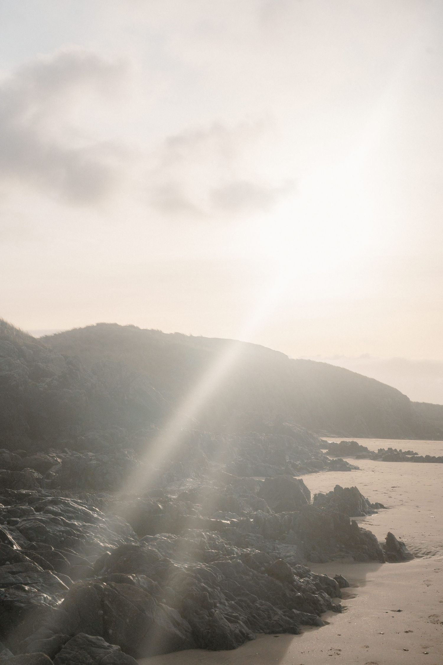 Misty coastal landscape with rocky shoreline, sandy beach, and dramatic sun rays breaking through hazy clouds over hills.