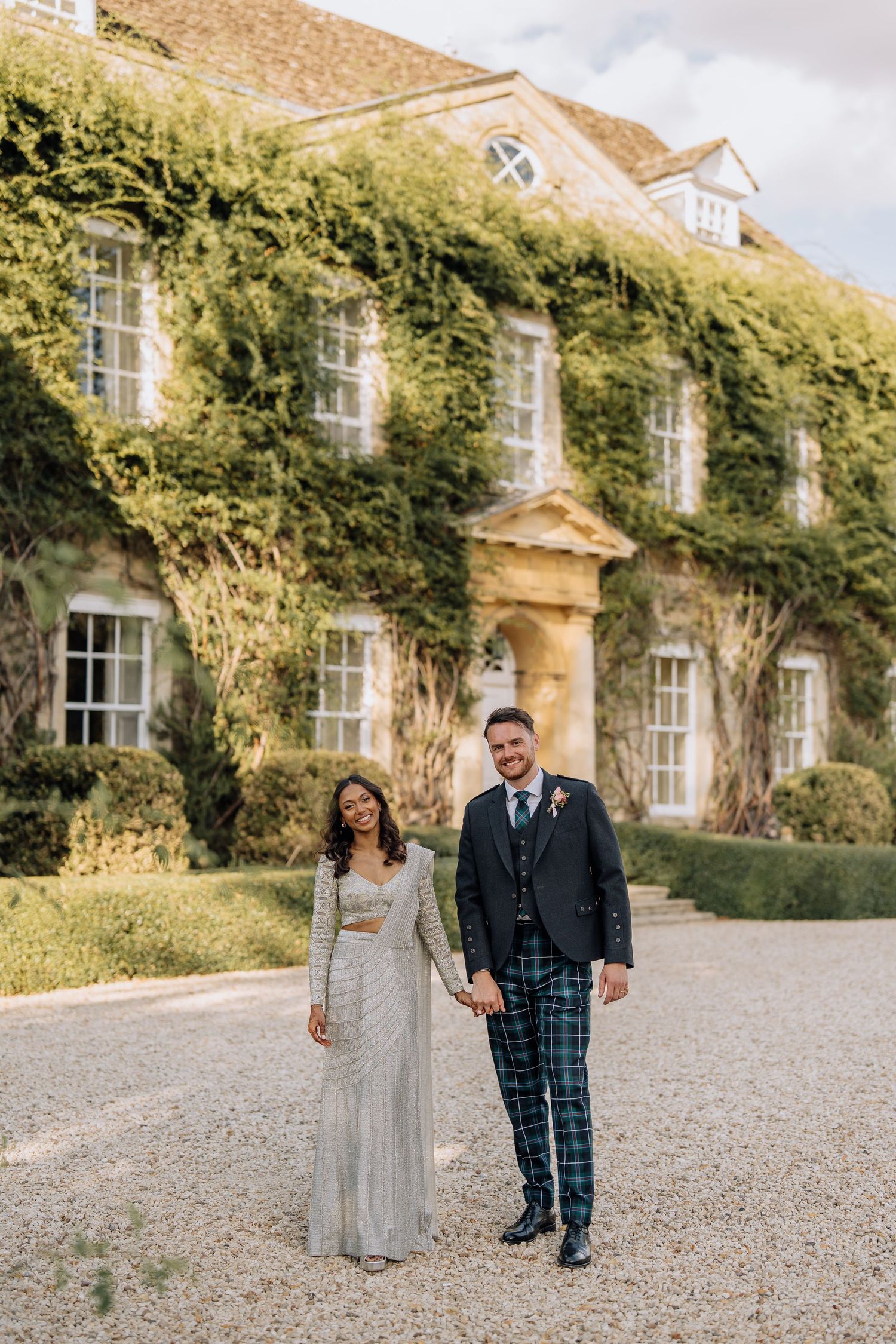 Interracial couple holding hands outside ivy-covered manor house, bride in silver gown and groom in tartan trousers with navy blazer.