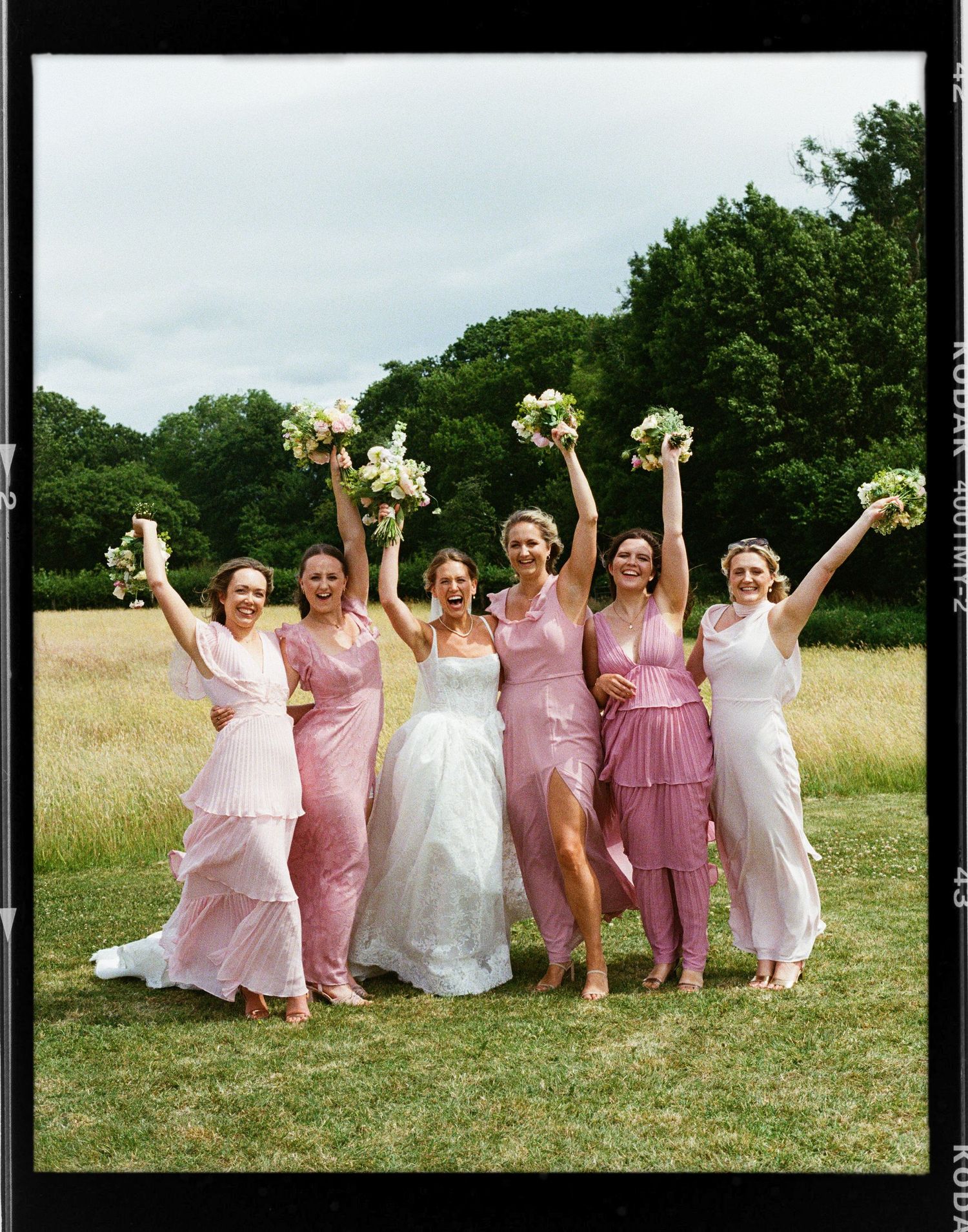 Bride in white gown with five bridesmaids in pink dresses raising bouquets outdoors in sunny field.