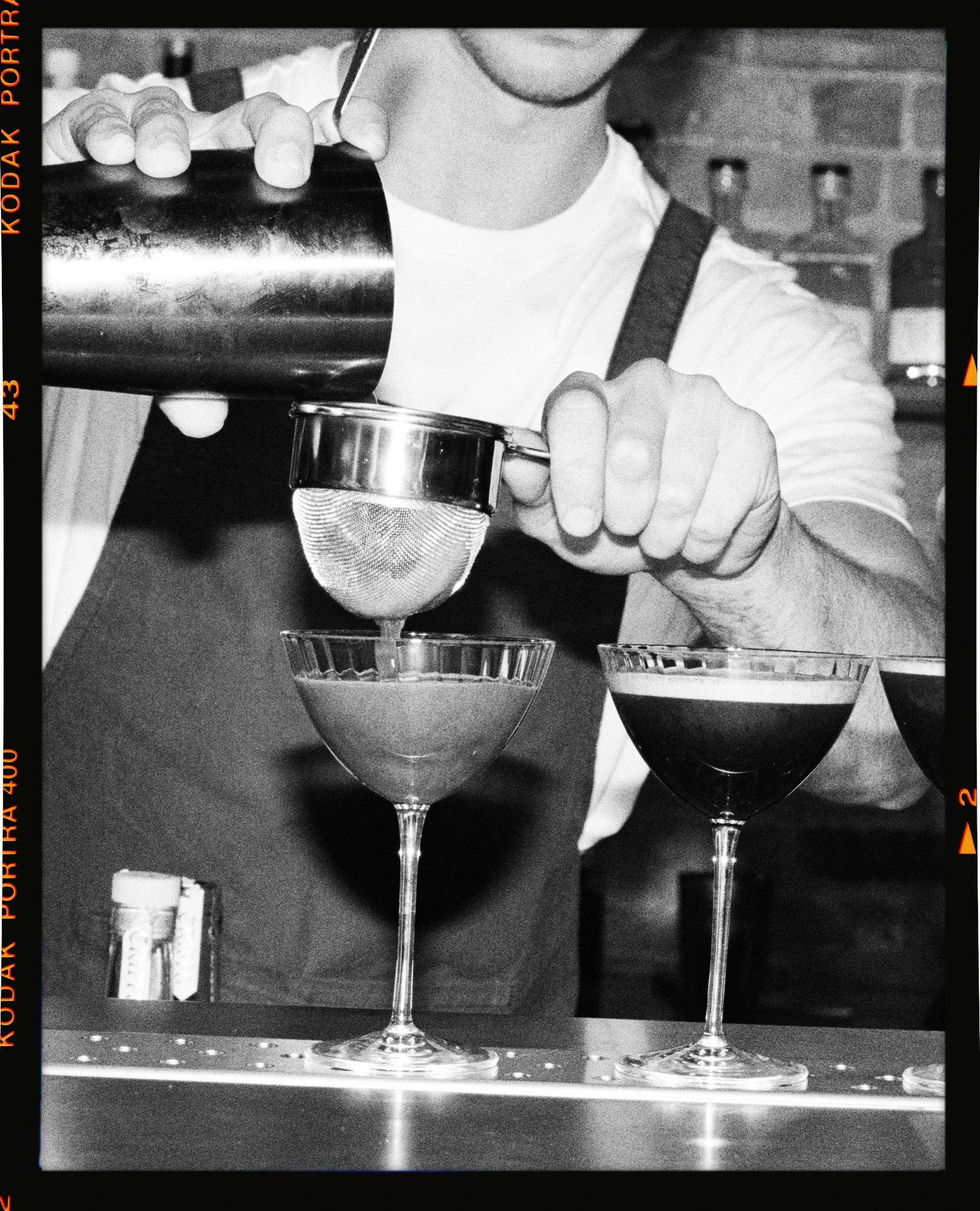 Bartender in black apron pouring cocktail shaker into two espresso martini glasses, black and white film photography.