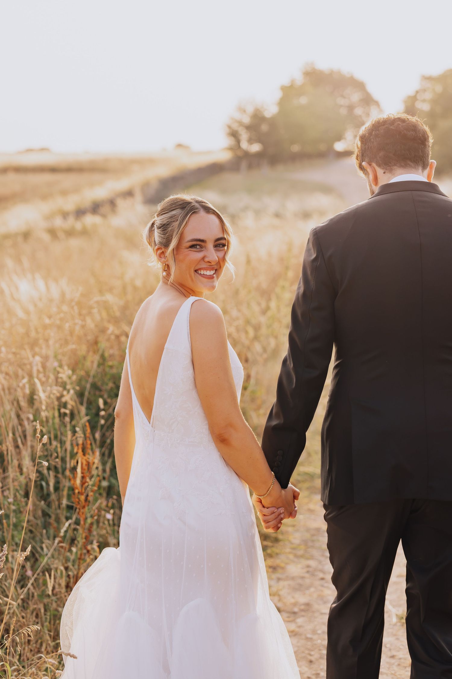 Bride in white gown smiles over shoulder holding groom's hand in golden sunset field.