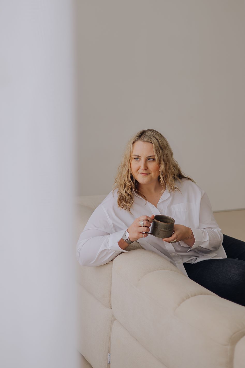 Blonde woman in white shirt holding a ceramic mug while relaxing on a white sofa.