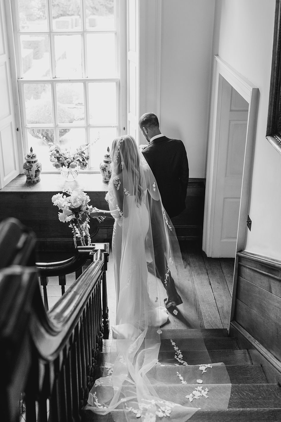 Black and white overhead view of bride in embroidered cathedral veil and groom descending grand staircase together.