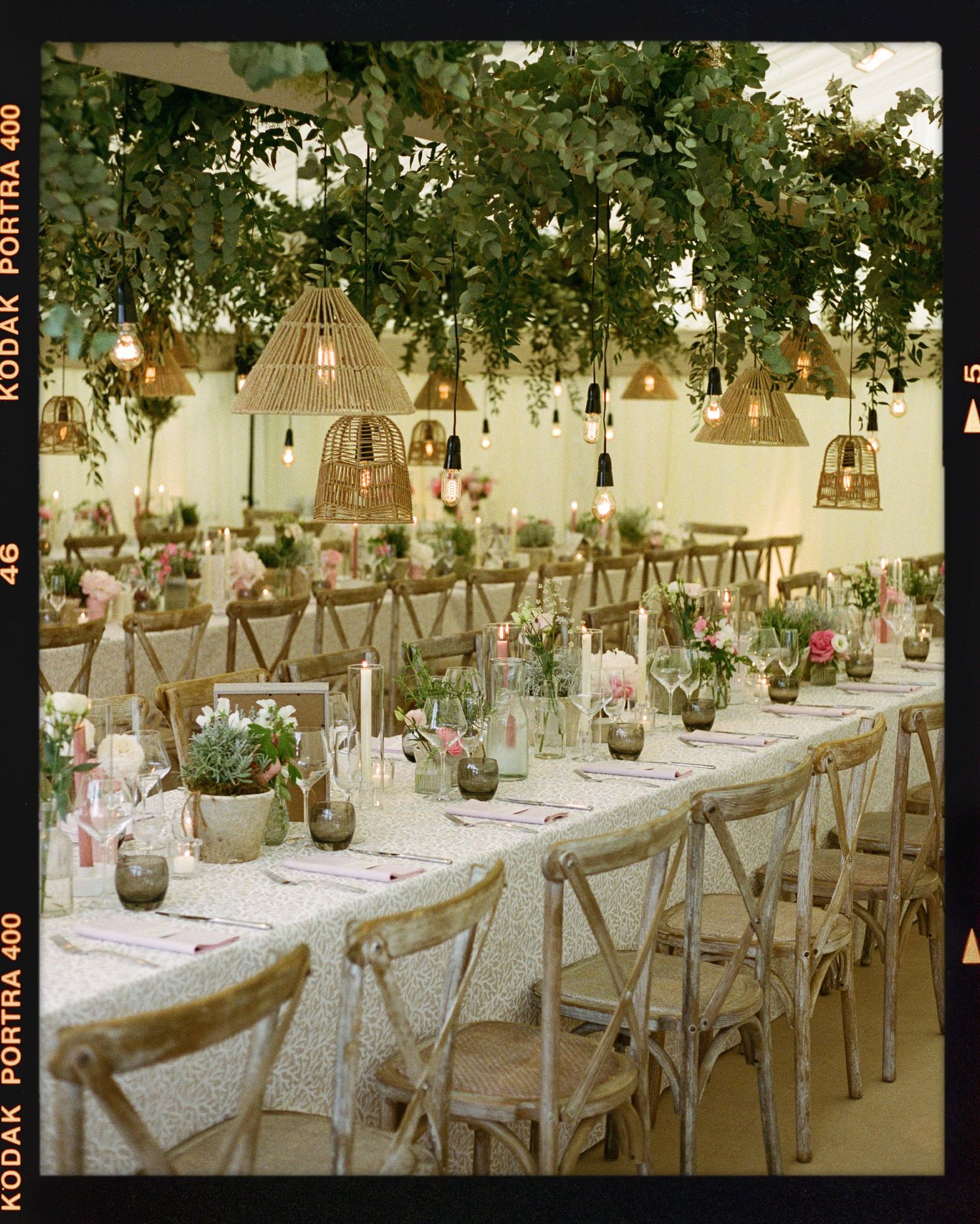 Elegant wedding reception with rustic wooden chairs, floral centerpieces, wicker pendant lights, and lush greenery canopy overhead.