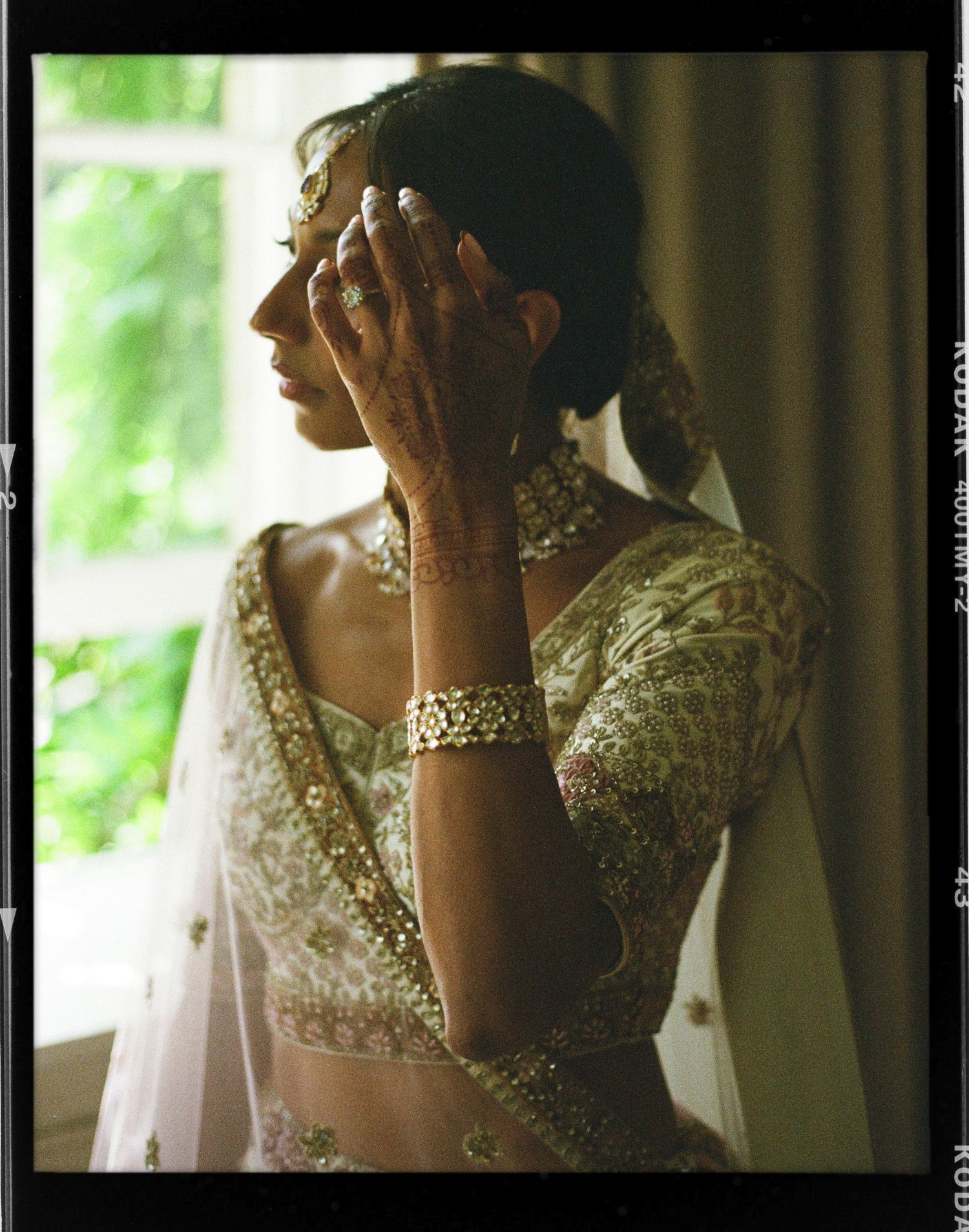 South Asian bride in gold embroidered lehenga with jewelry and mehndi poses by window light.