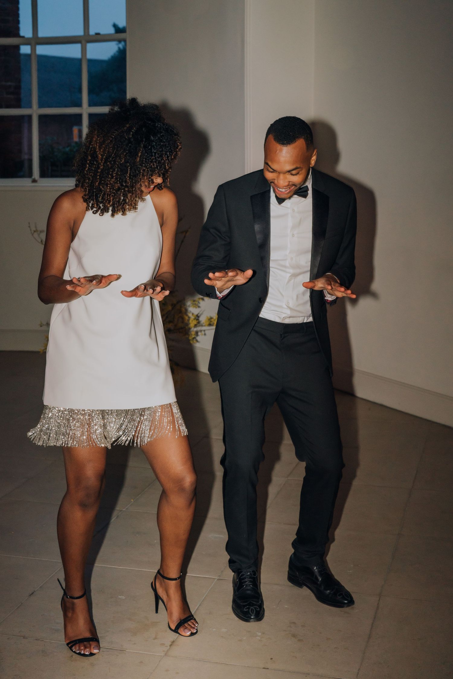 Bride in white fringe mini dress and groom in tuxedo share energetic dance moves at wedding reception.