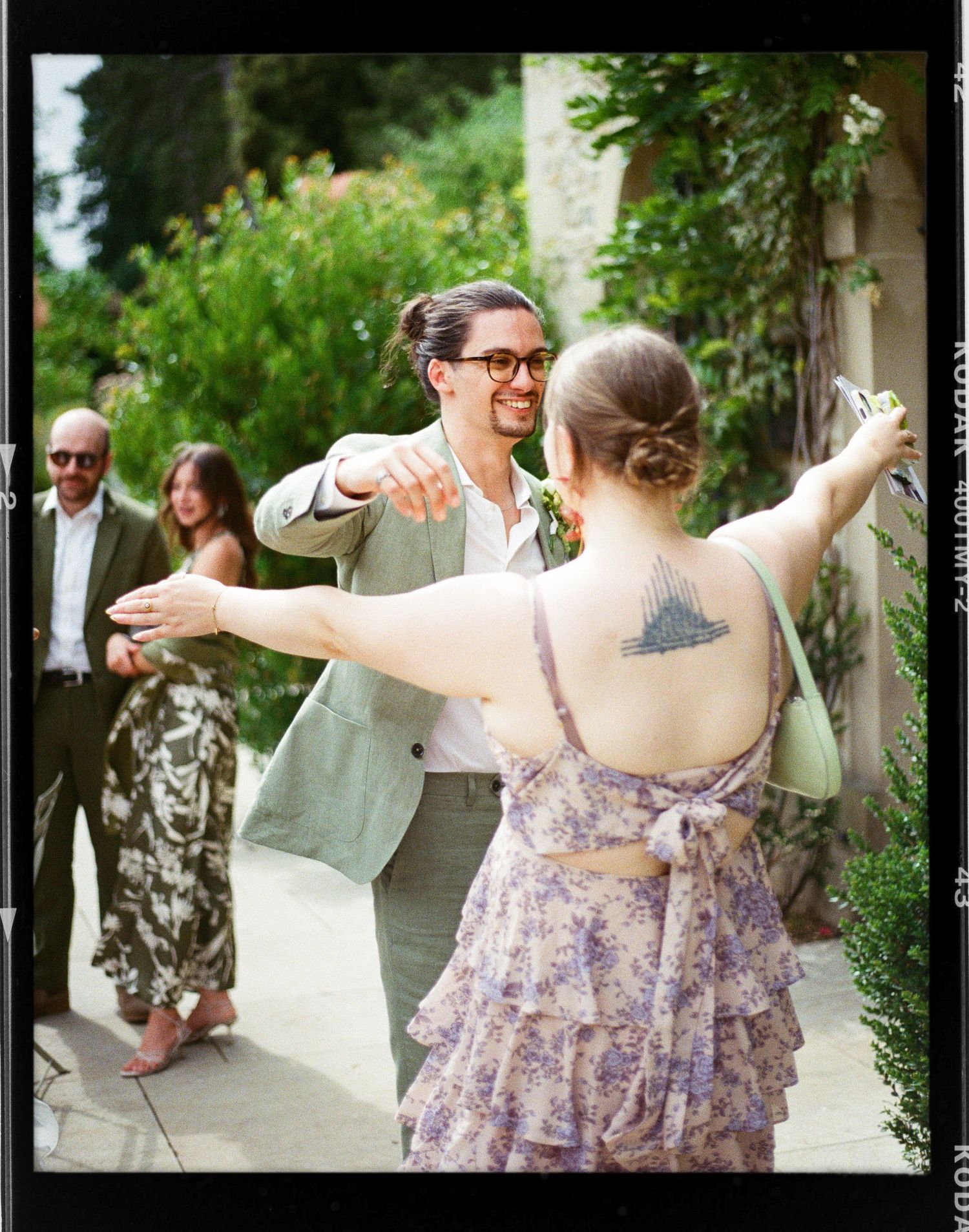 Man in green suit dancing with tattooed woman in floral dress at outdoor garden party.