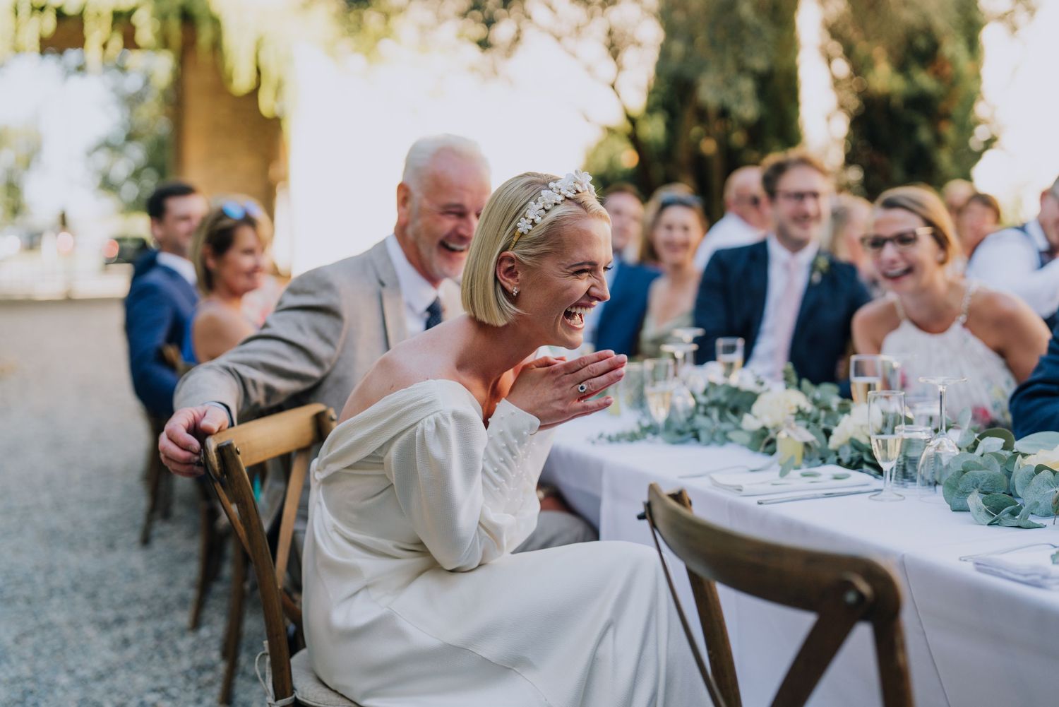 Bride laughing joyfully at wedding reception dinner table surrounded by smiling guests in outdoor setting.