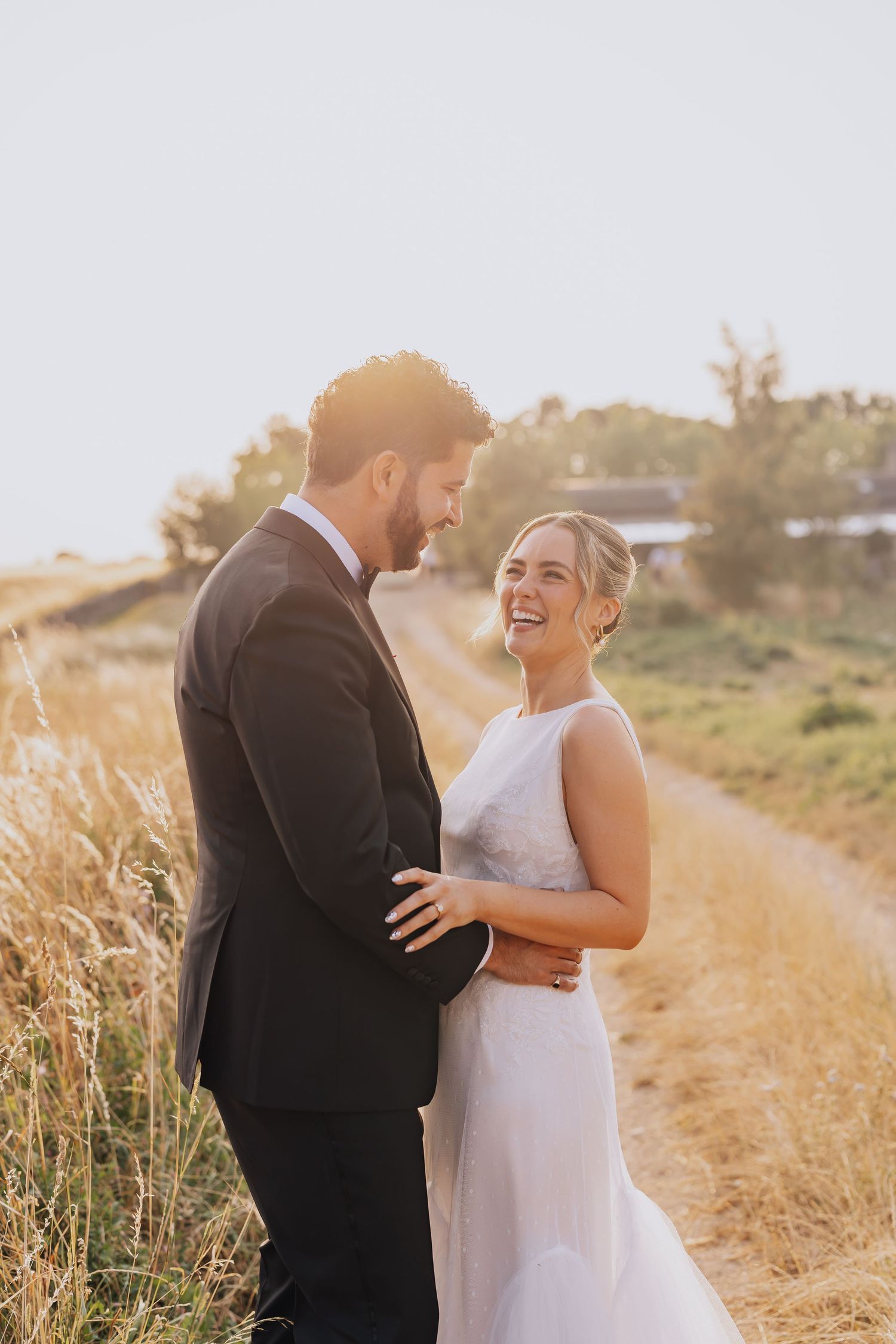 Bride and groom share a joyful moment on a golden field path during sunset in wedding attire.